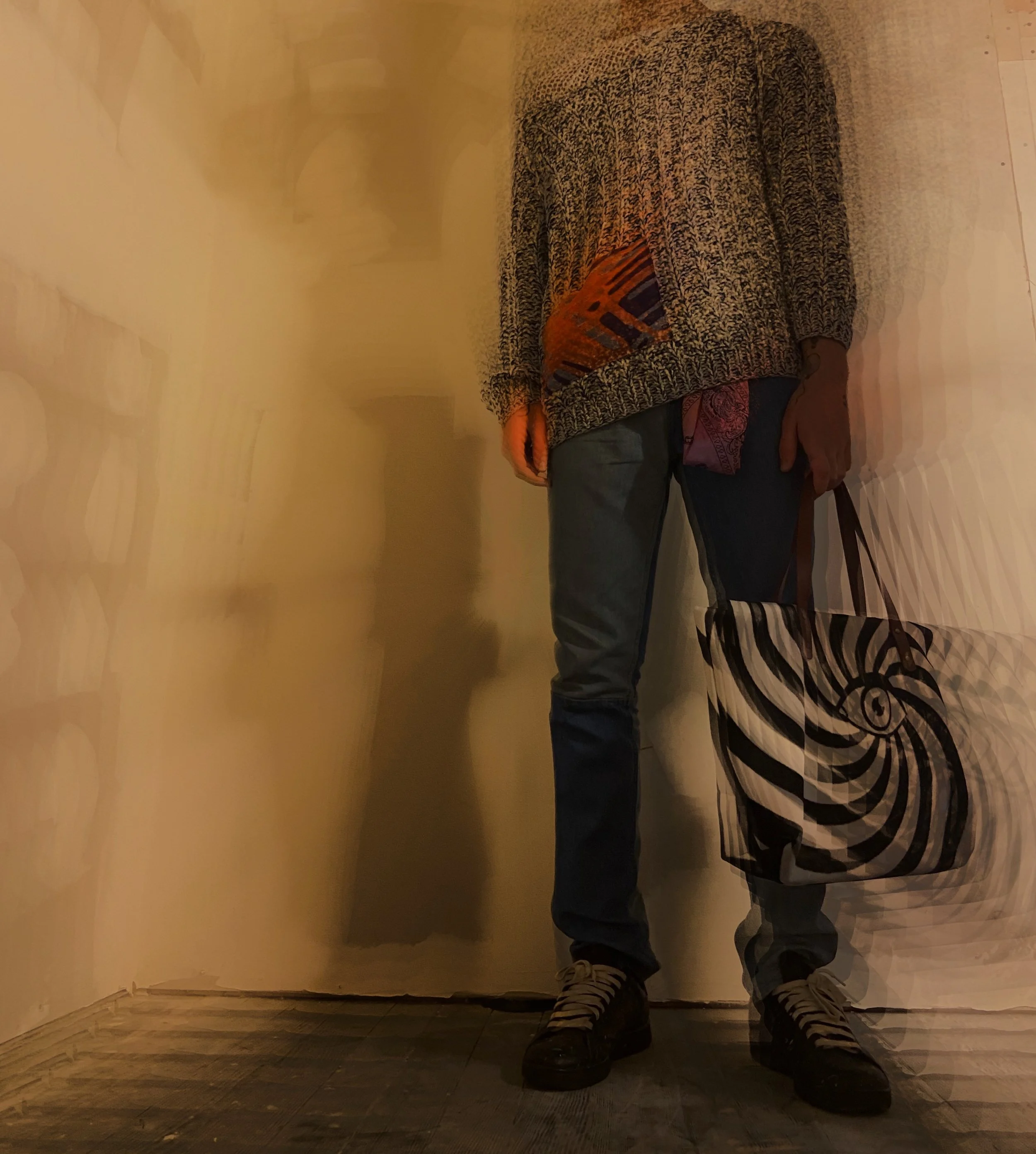 Person standing against a wall, wearing a patterned sweater, jeans, and holding a black and white spiral patterned tote bag.
