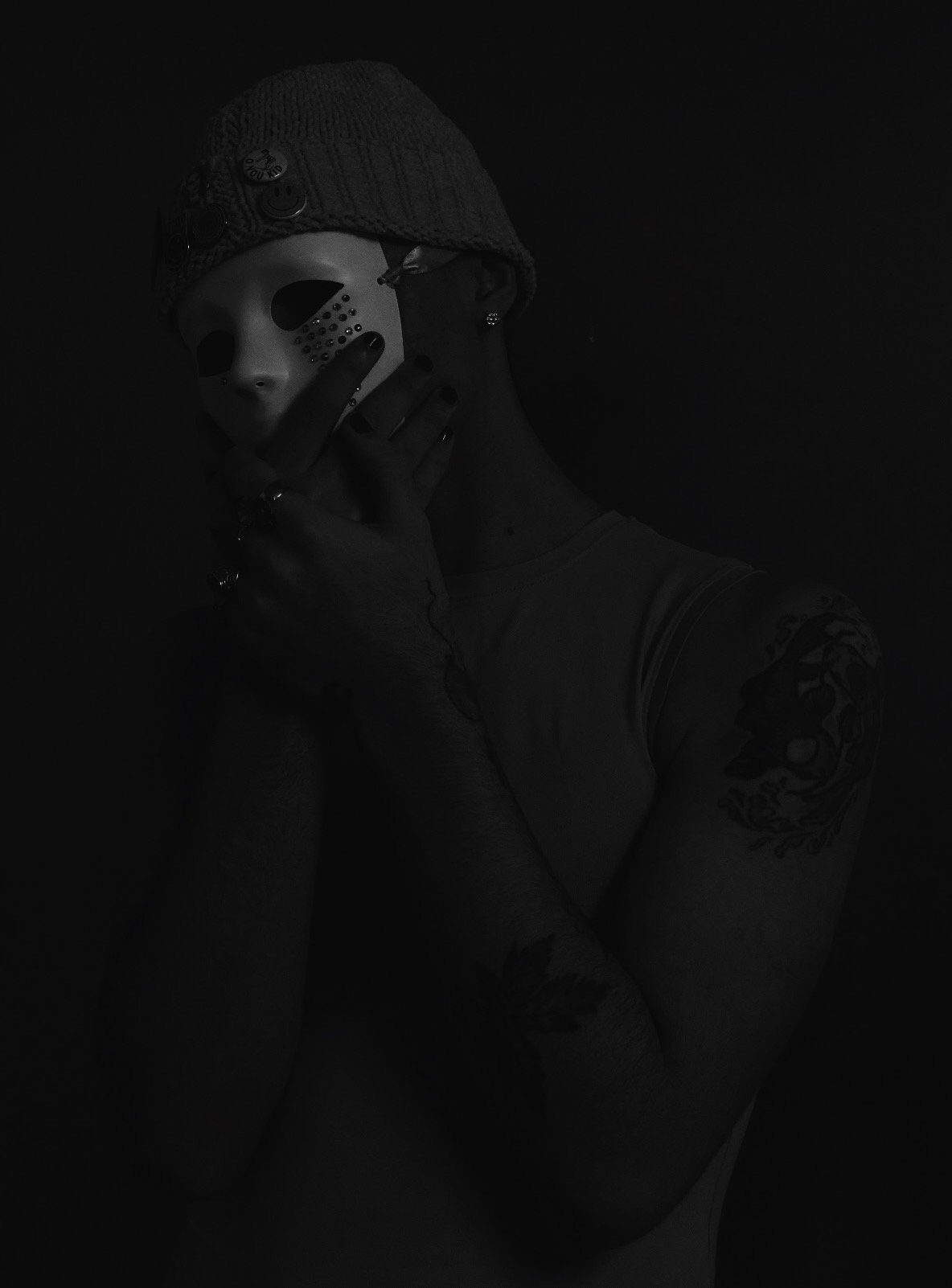 Person wearing a knit beanie, holding a hockey mask in front of their face, with tattoos visible on their arm and shoulder, in a dark setting.