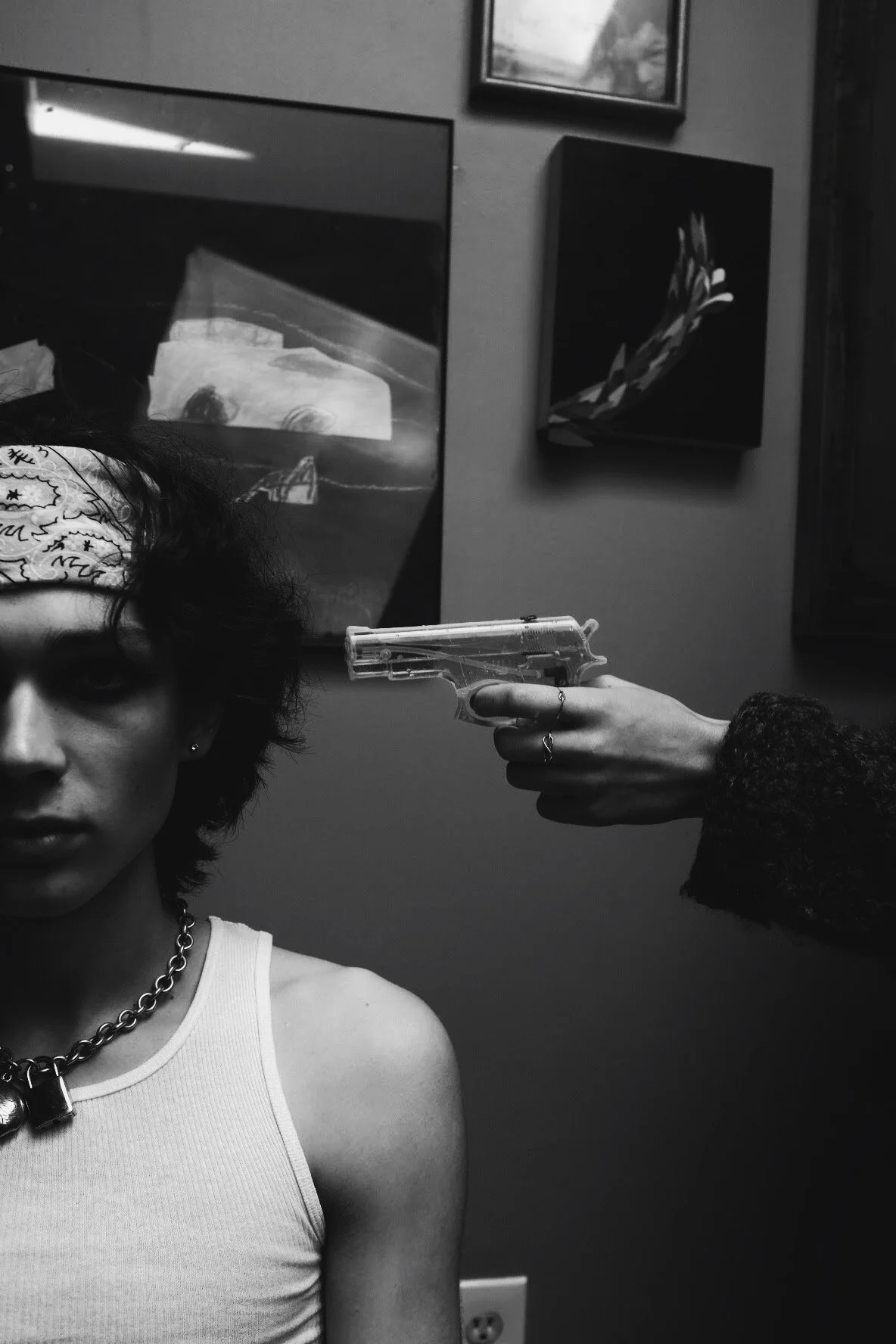Black and white photo of a woman sitting with a bandana on her head, wearing a sleeveless top and a chain necklace, while another person holds a gun to her head from behind.