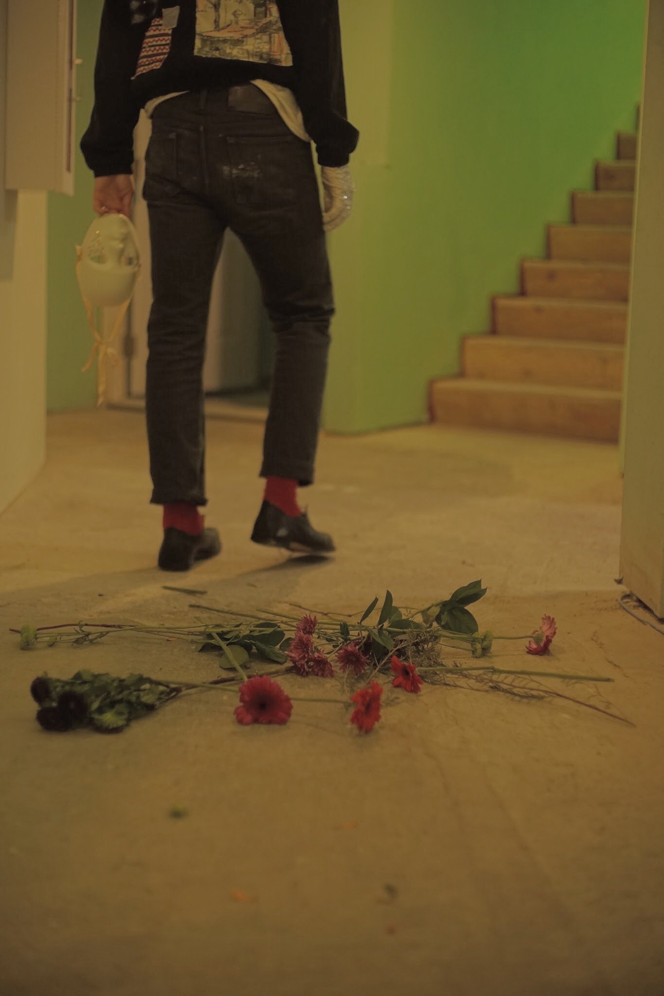 A person standing in a hallway near a staircase, holding a pair of shoes, with wilted flowers lying on the floor.