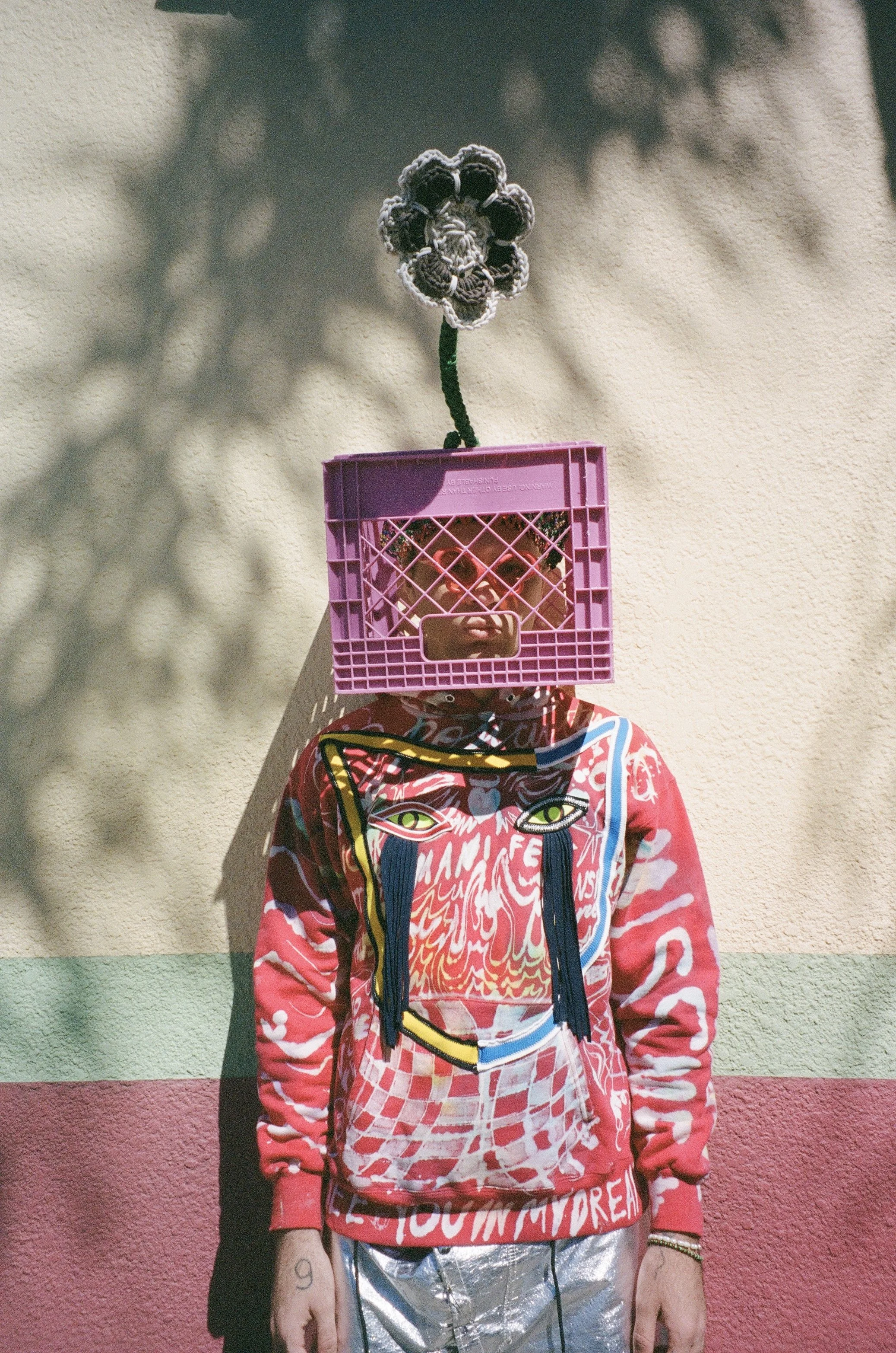 Person wearing a colorful hoodie with a graphic design, shiny silver pants, and a pink plastic crate on their head with a crocheted flower attached, standing against a wall with shadow patterns.