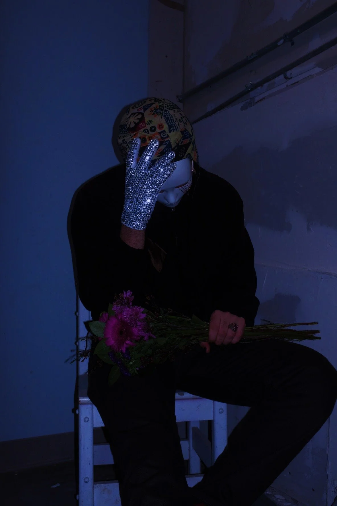 Person wearing a colorful headscarf, a white mask, and sparkly gloves, holding a bouquet of purple flowers, sitting on a white chair in a dimly lit room.