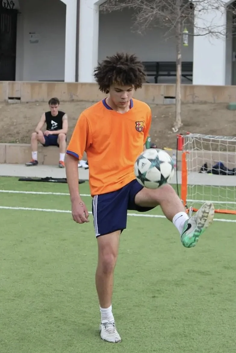 Young male soccer player practicing on a field, wearing an orange sports jersey and navy shorts, controlling a soccer ball with his foot.