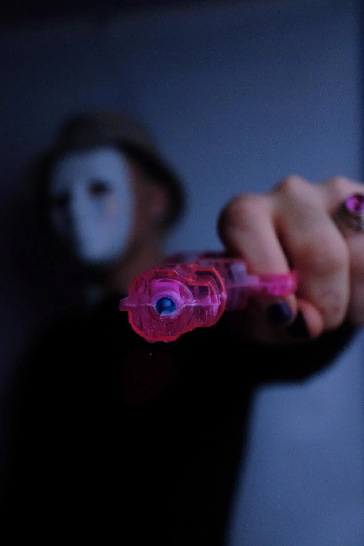 A person wearing a white mask and black outfit holds a pink toy gun pointed towards the camera in a dark room.