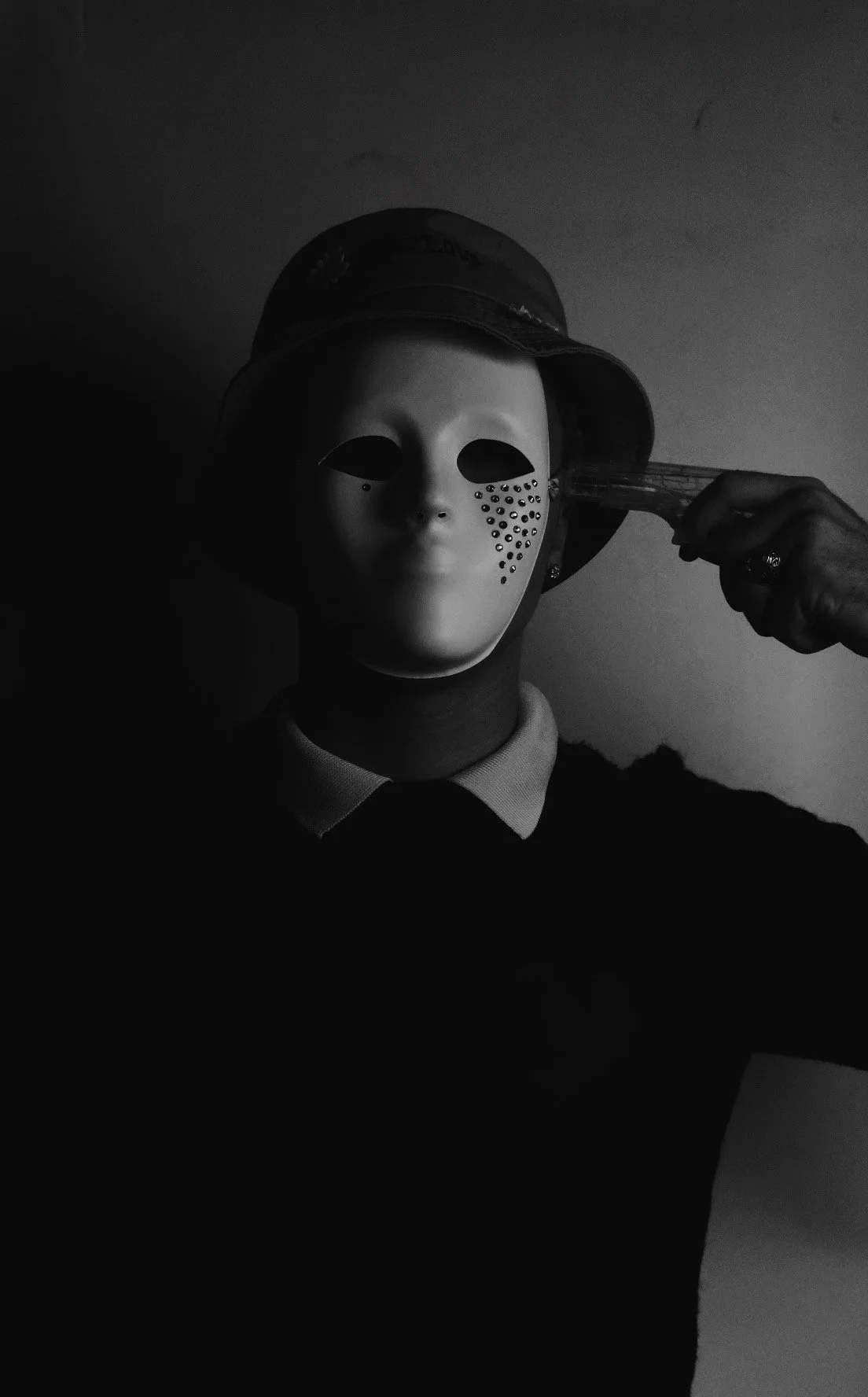 Person wearing a mask with dotted pattern on the face, holding a gun to their head wearing a bucket hat in a dark setting.