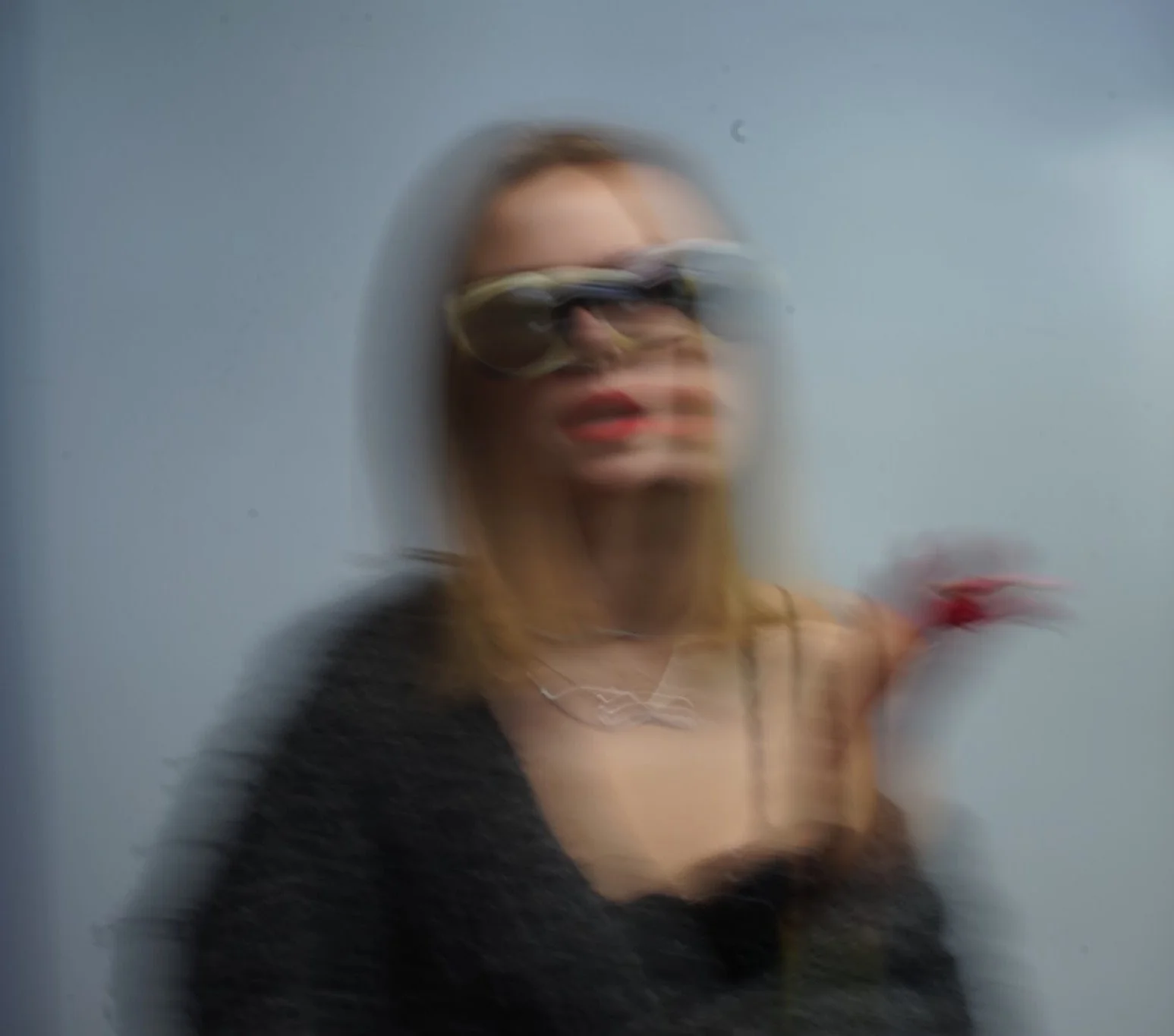 Blurred image of a woman wearing glasses and a black top, with light-colored hair, standing against a blue background.