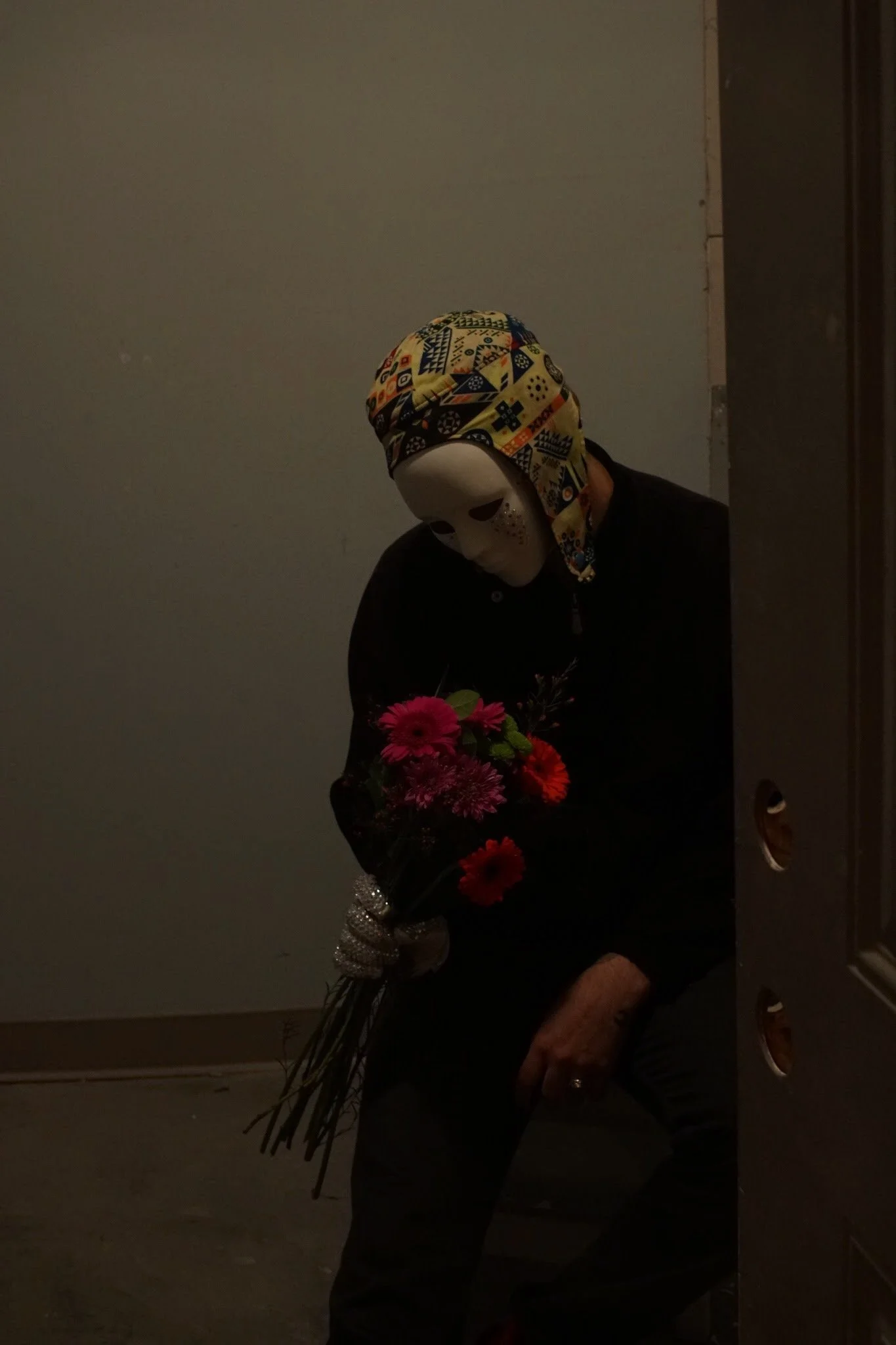 Person wearing a white mask with a colorful headscarf, holding a bouquet of red and pink flowers, standing in a dimly lit room.
