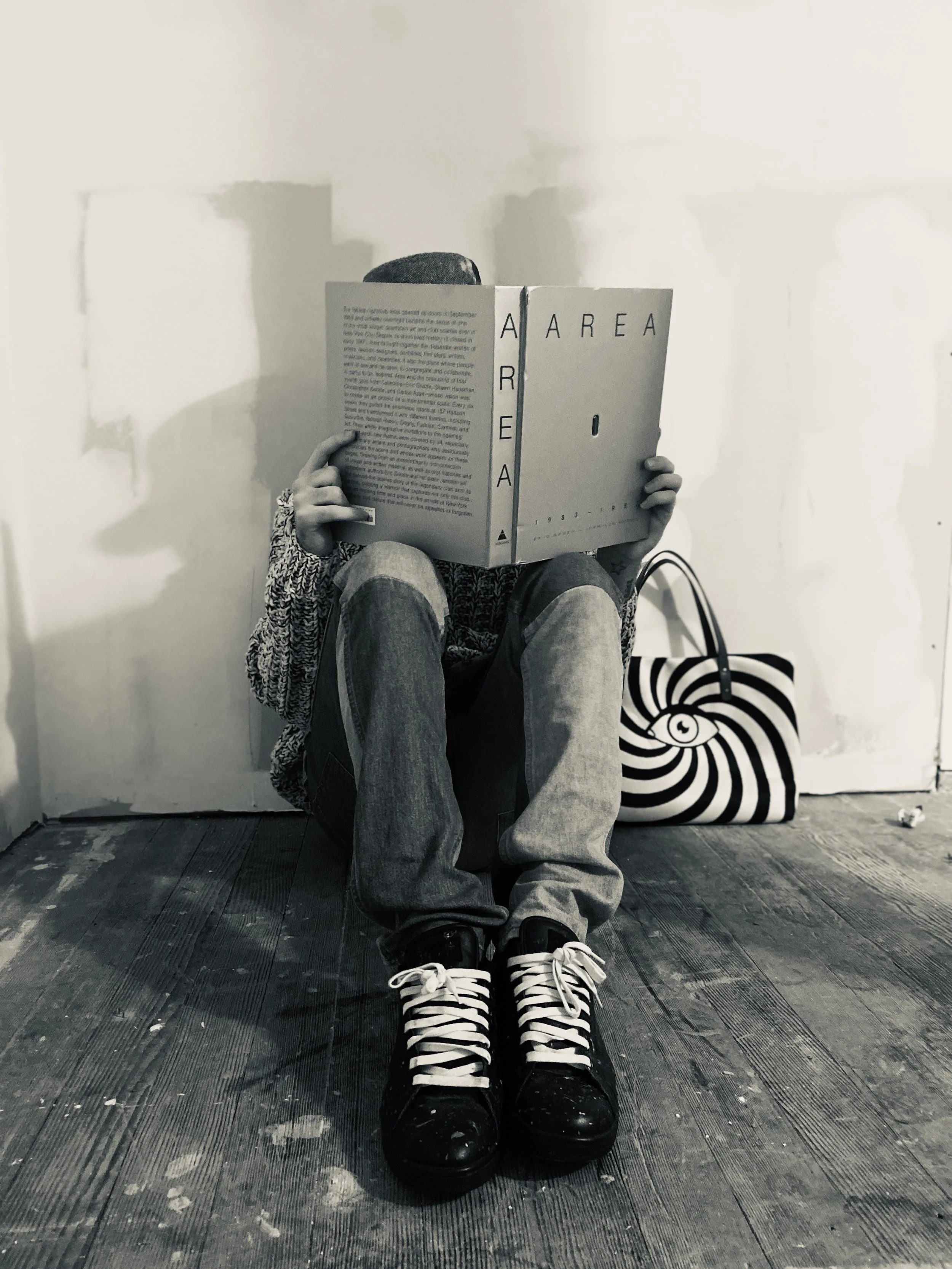 A person sitting on the floor reading a large magazine or book, with a striped bag beside them. They wear sneakers, jeans, and a patterned sweater. The background is plain with some patches on the wall, and the floor appears to be wooden.