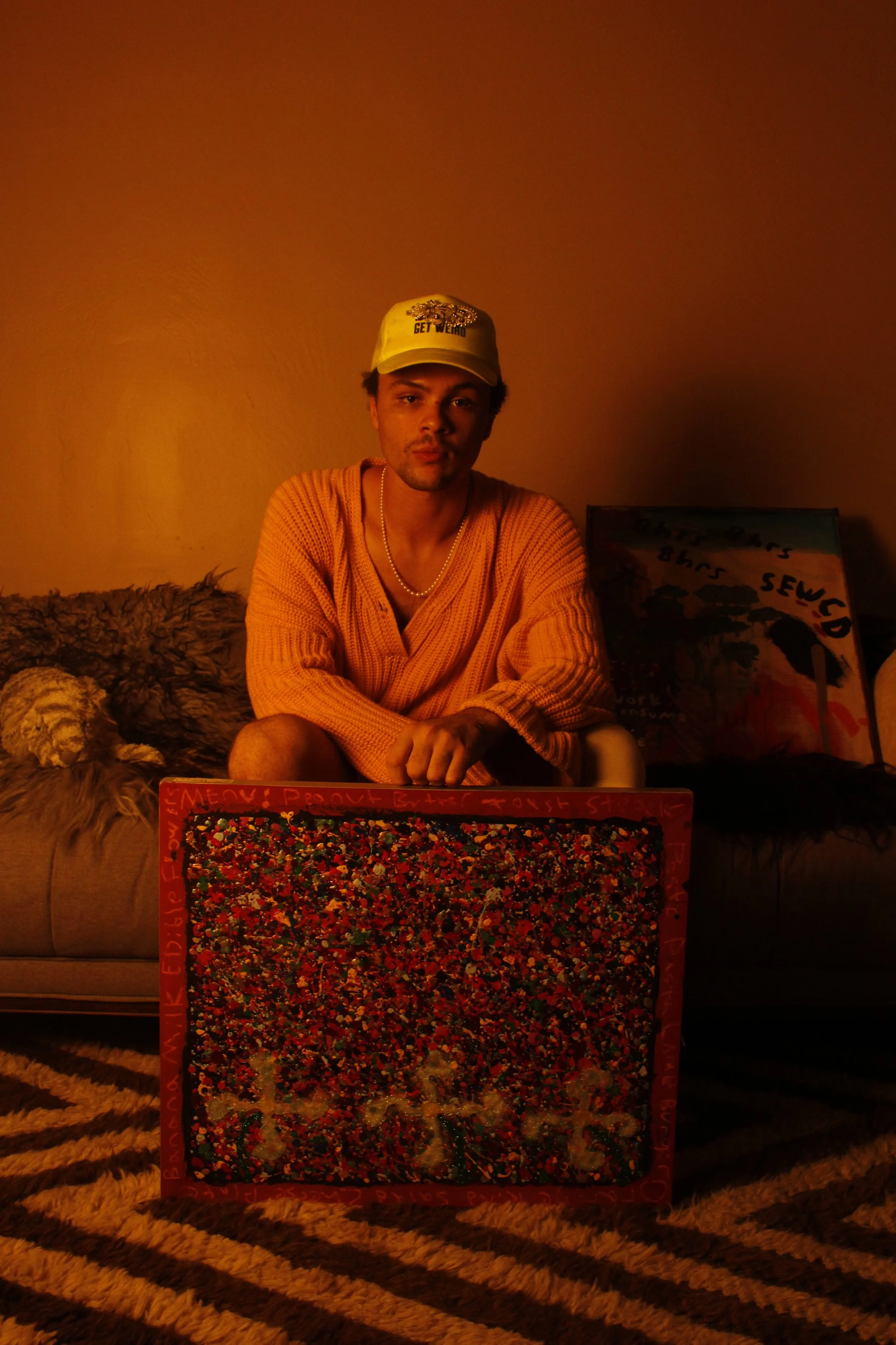 A young man sitting on a couch in a warmly lit room, wearing a yellow cap and orange sweater, with artwork behind him and a colorful painting in front.