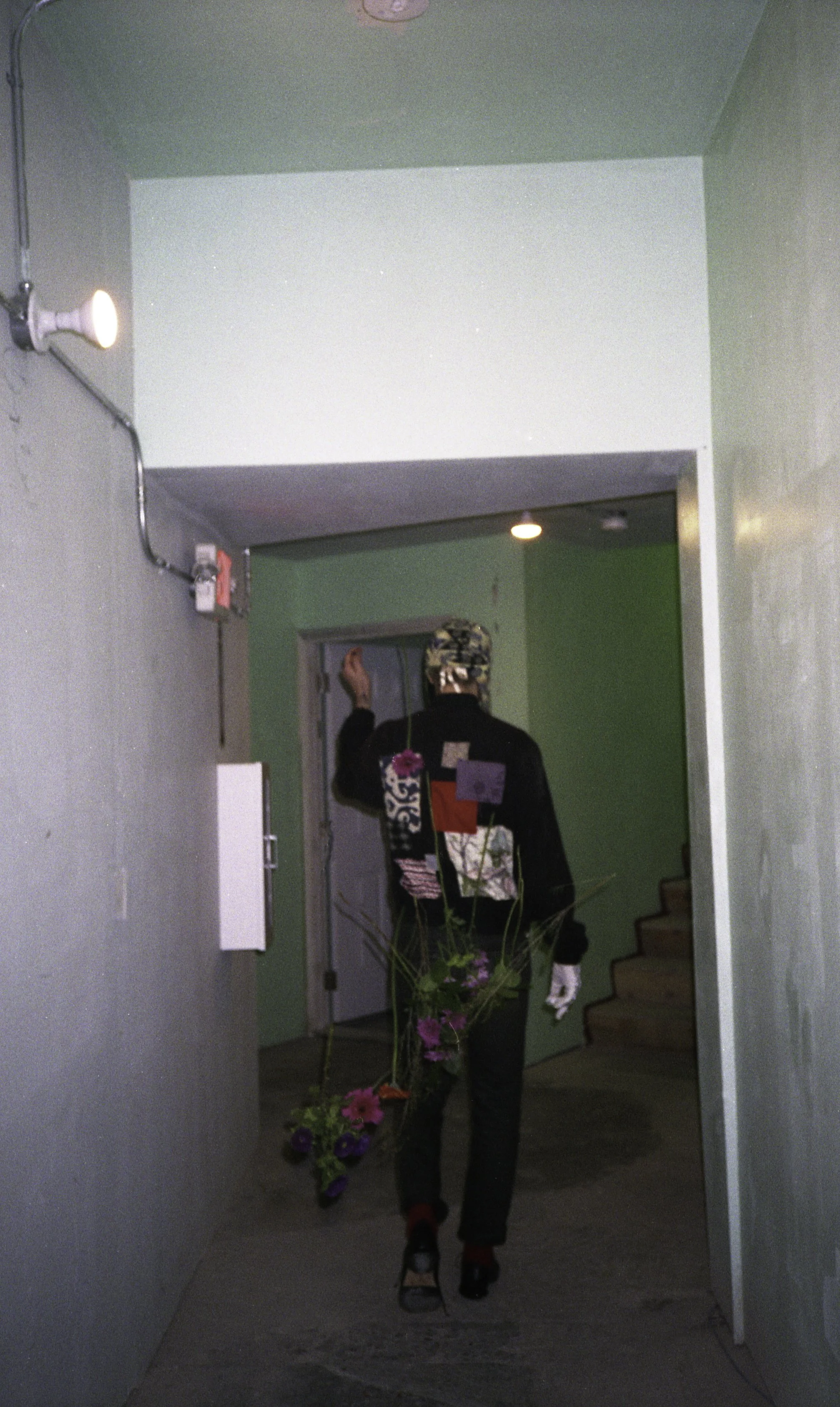 A person with a hat, patterned jacket, and black pants holding a bouquet of pink and purple flowers, walking into a building within a hallway with green walls.
