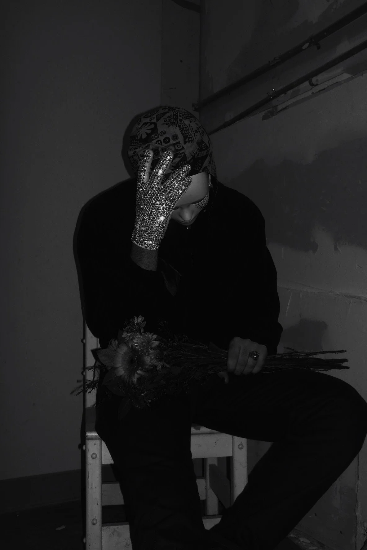 A person wearing a patterned headscarf and sparkling glove, holding a bouquet of flowers, sitting on a chair in a dimly lit room with an industrial-style wall.