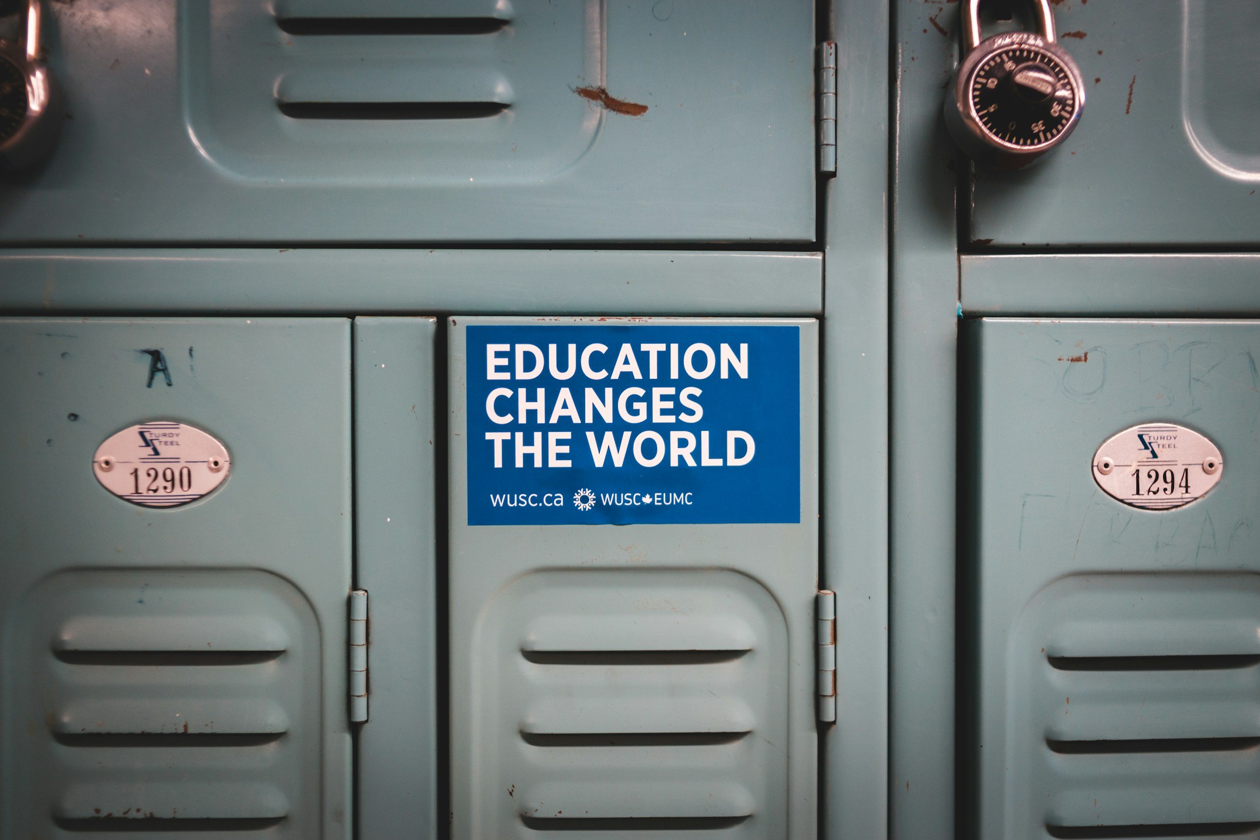A row of old metal lockers painted light blue, with rust spots and scratches. A blue sticker on one locker reads 'Education Changes the World' with website and organization logos.