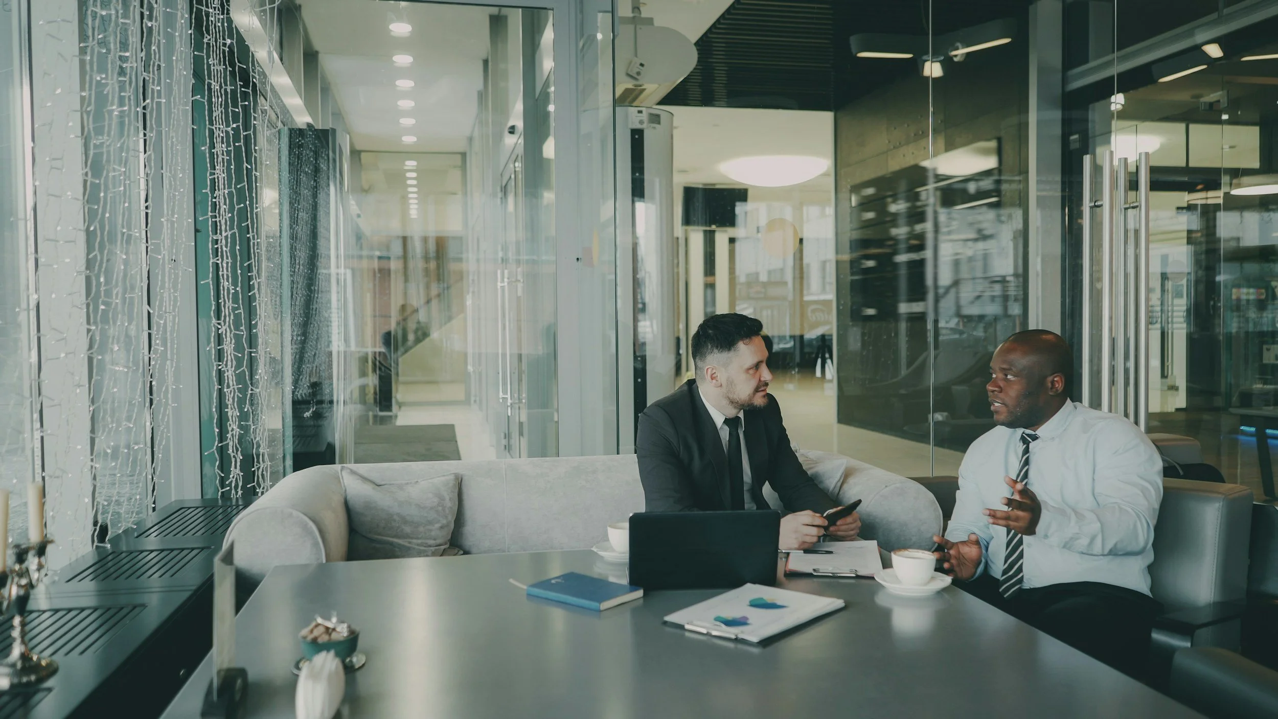 Two men in business attire conversing in a modern office lobby with glass walls and string lights, seated at a table with coffee cups, a laptop, a notebook, and papers.