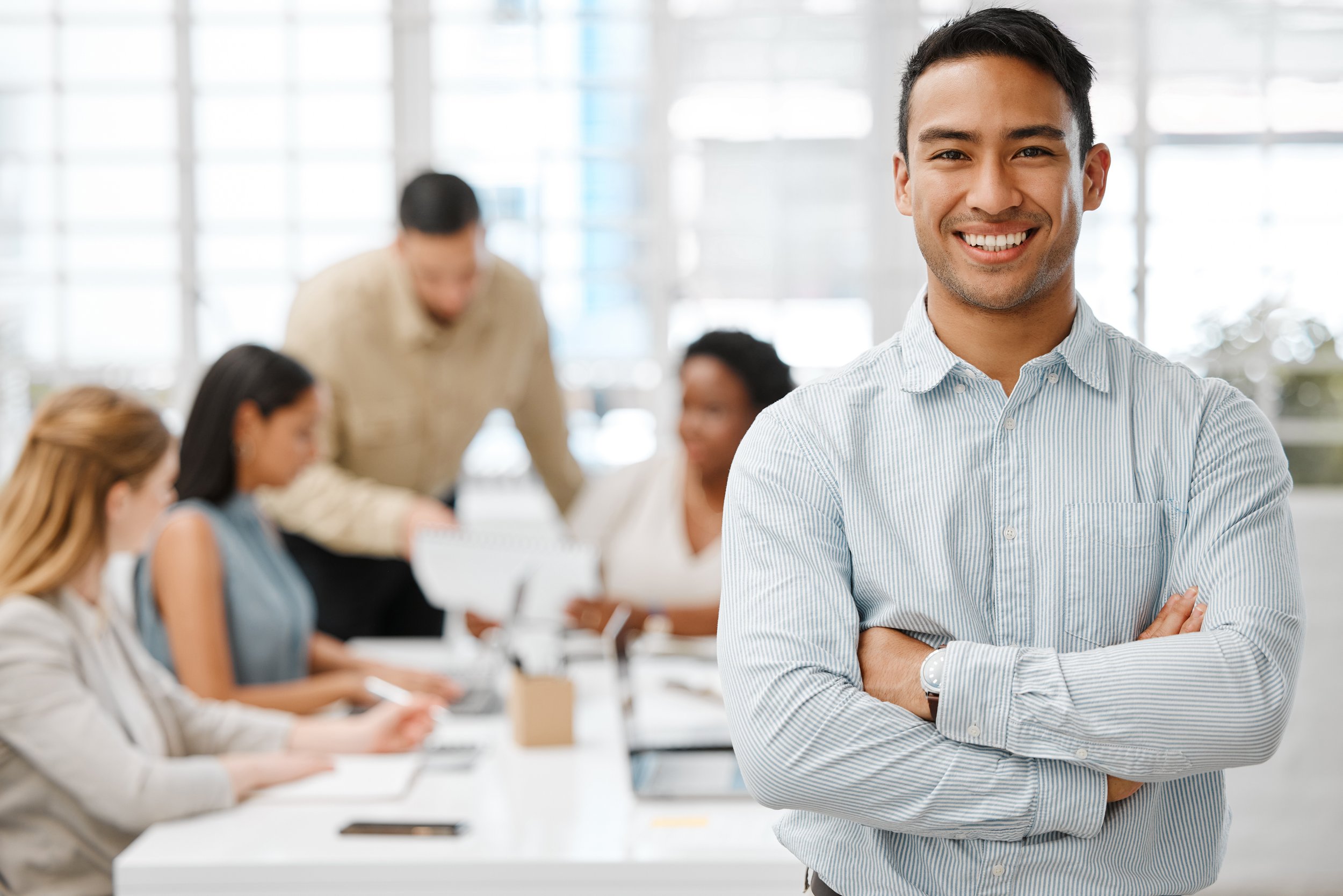 A young man with dark hair, smiling, wearing a light blue striped shirt, standing with arms crossed in a bright office space with large windows. In the background, a group of diverse women are seated at a table, engaged in a discussion or working on laptops.