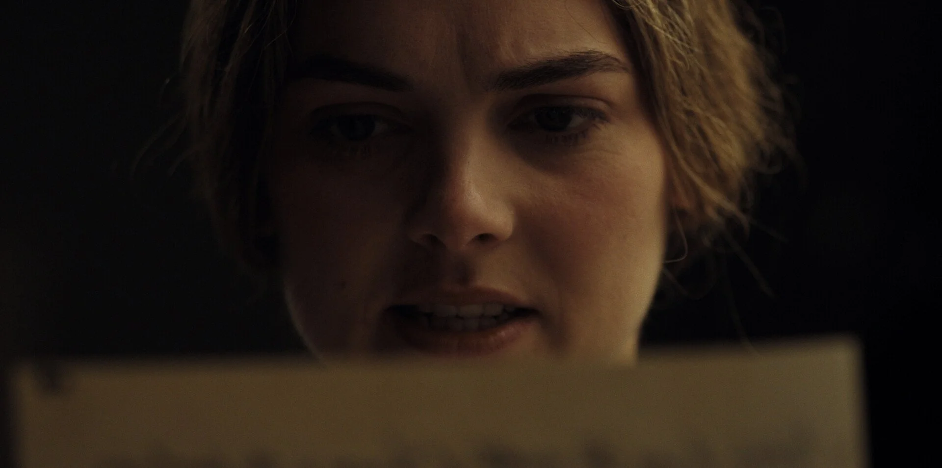 Rose Riley in close-up, intently reading a document with a serious expression in dim lighting in The Mark (2025)