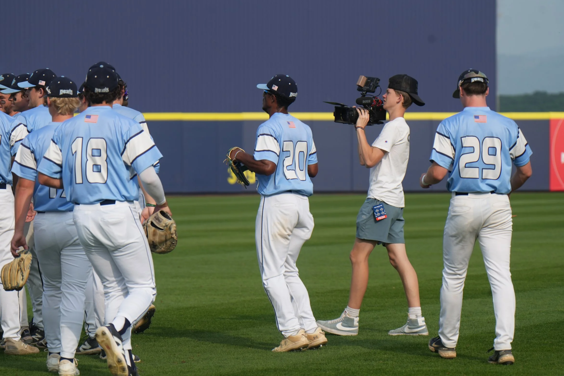 Baseball players in light blue jerseys and white pants gather on a baseball field, with one player having a glove, and a woman filming them with a video camera.