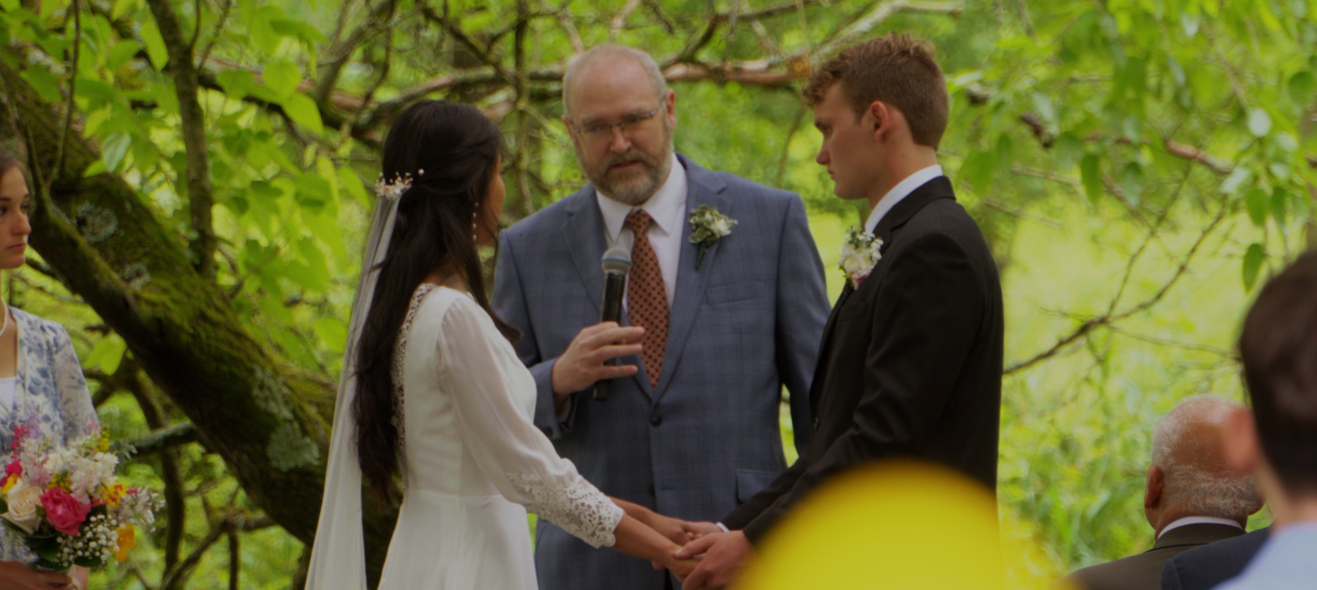 A couple getting married under green trees, holding hands, with an officiant and attendees around, during an outdoor wedding ceremony.