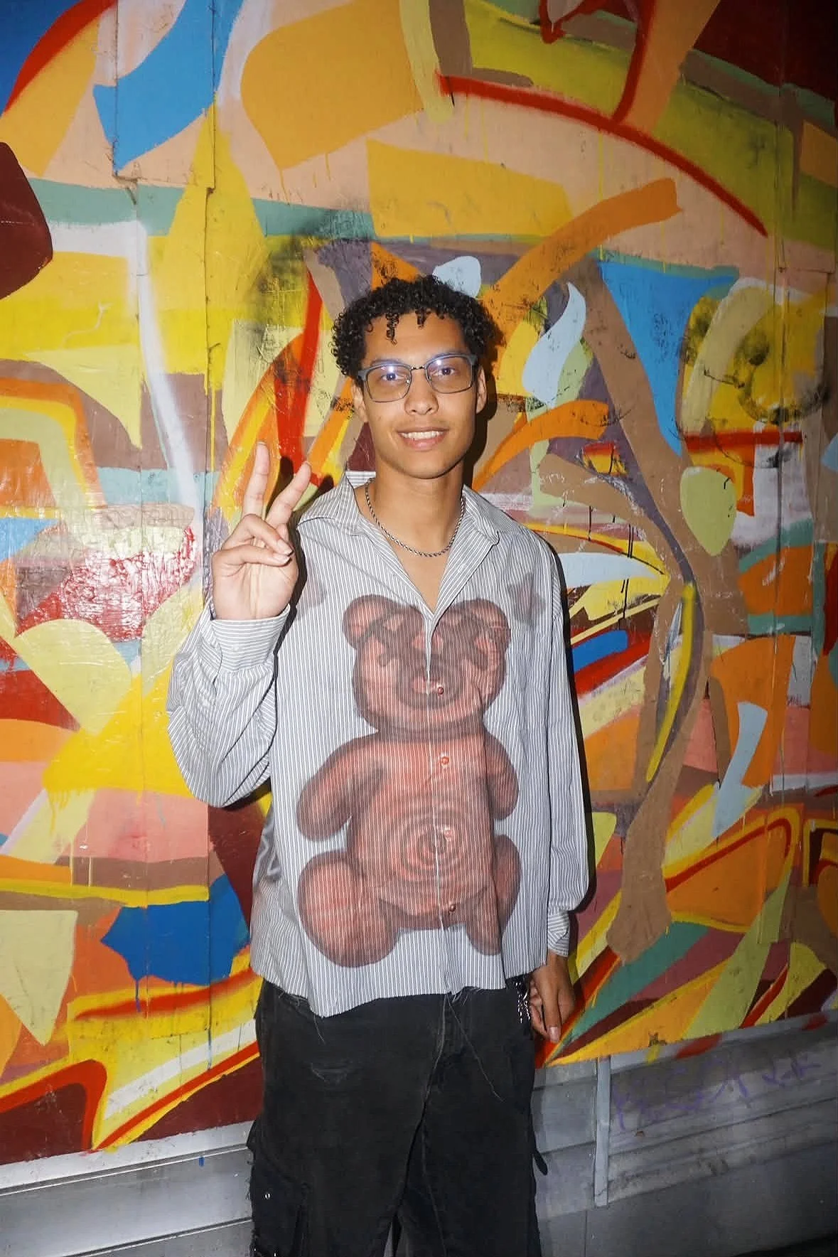 Jalen Jordan with curly hair and glasses making a peace sign with his right hand, standing in front of a colorful abstract mural painting.