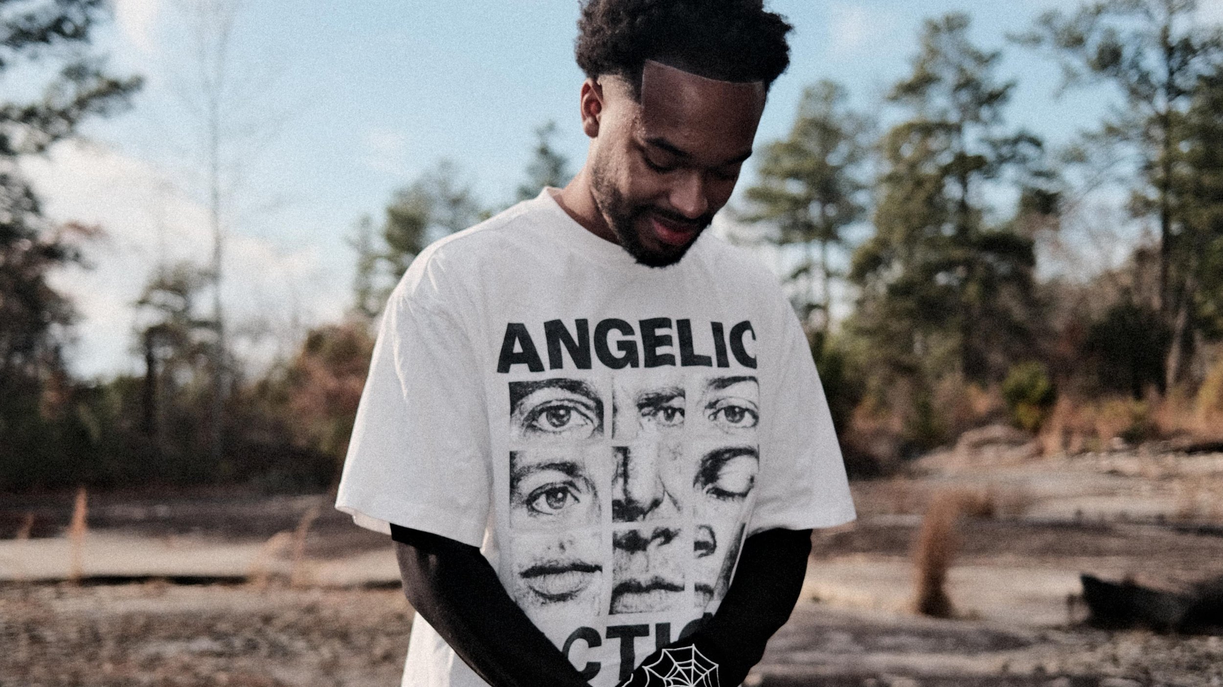 Jx.Zero wearing a white T-shirt with black graphic text and images stands outdoors on a cloudy day with trees in the background.