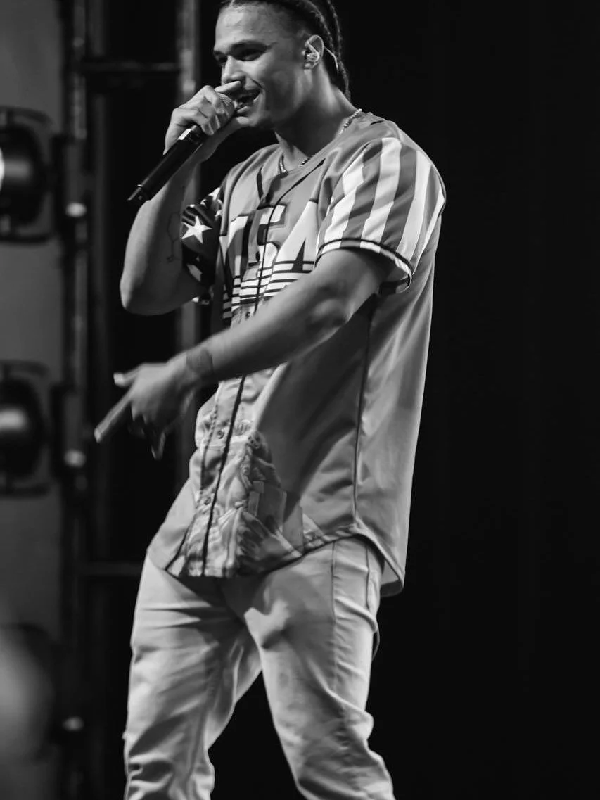 Deyton Jackson performing on stage, holding a microphone in his right hand, wearing a baseball jersey and light-colored pants, smiling with eyes closed in black and white photo.
