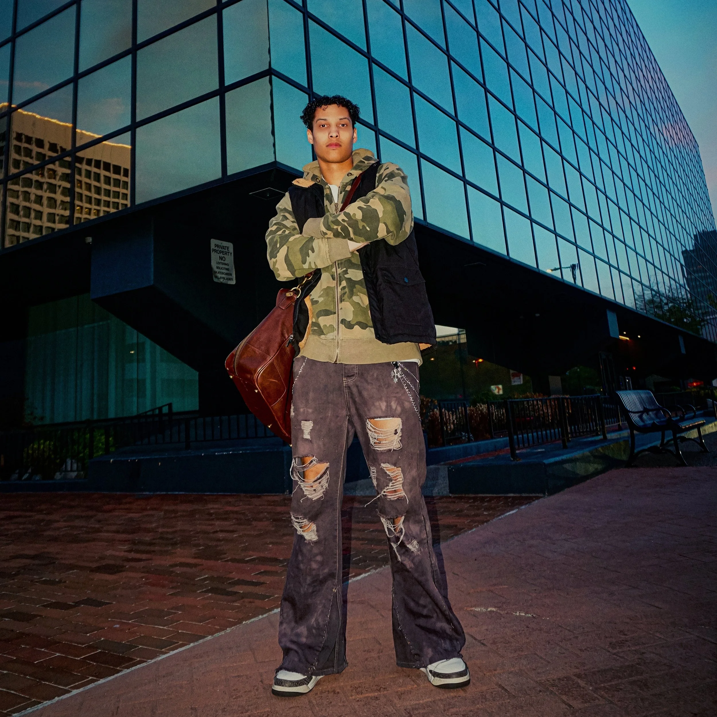 Jalen Jordan with curly hair wearing a camouflage hoodie and ripped jeans, standing with arms crossed in front of a modern glass building during the evening.