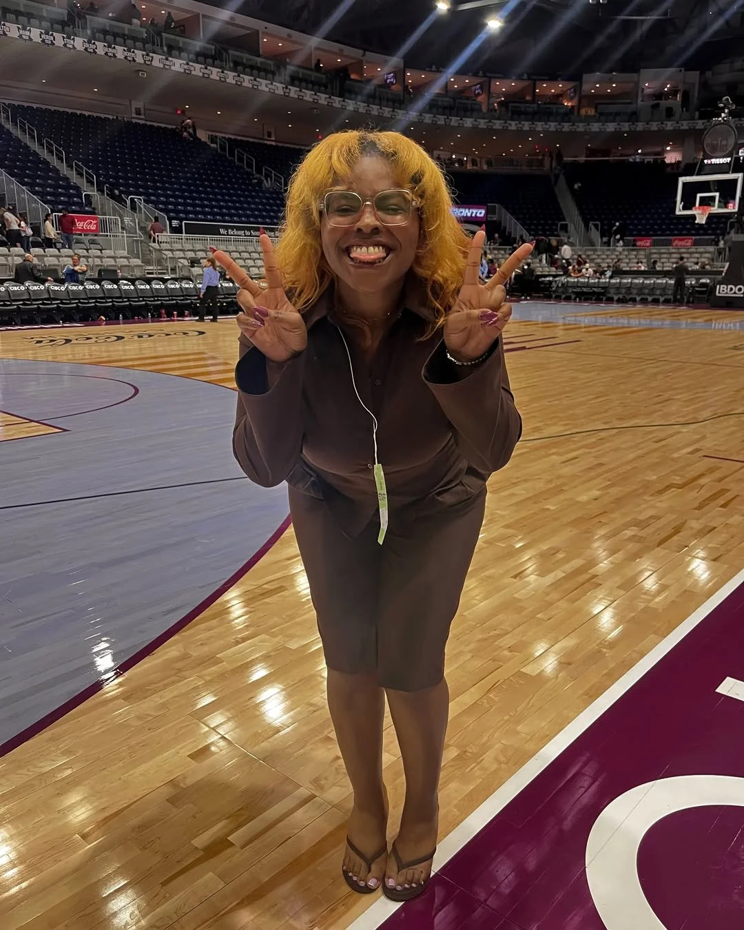  DĀHNI with blonde curly hair and glasses is standing on a basketball court, smiling and making peace signs with both hands.