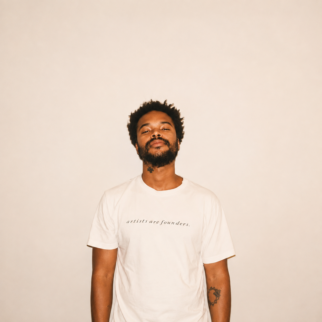 Jx.Zero with a beard and curly hair standing against a plain beige wall, wearing a white T-shirt with black text that reads "artists are founders."
