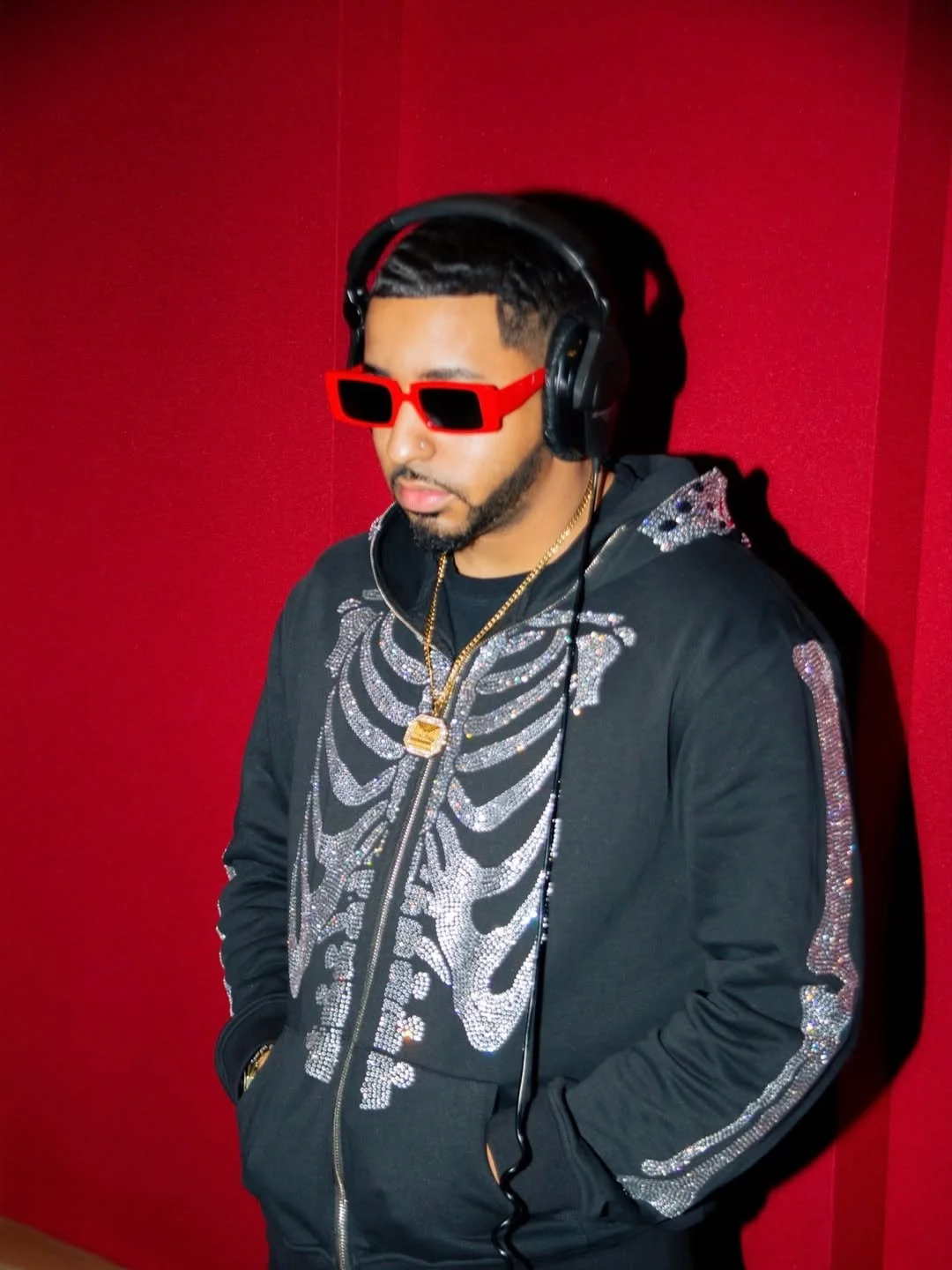 Fame Holiday wearing large red sunglasses, black headphones, a black jacket with silver rhinestone skeleton design, and gold jewelry, standing against a red wall.