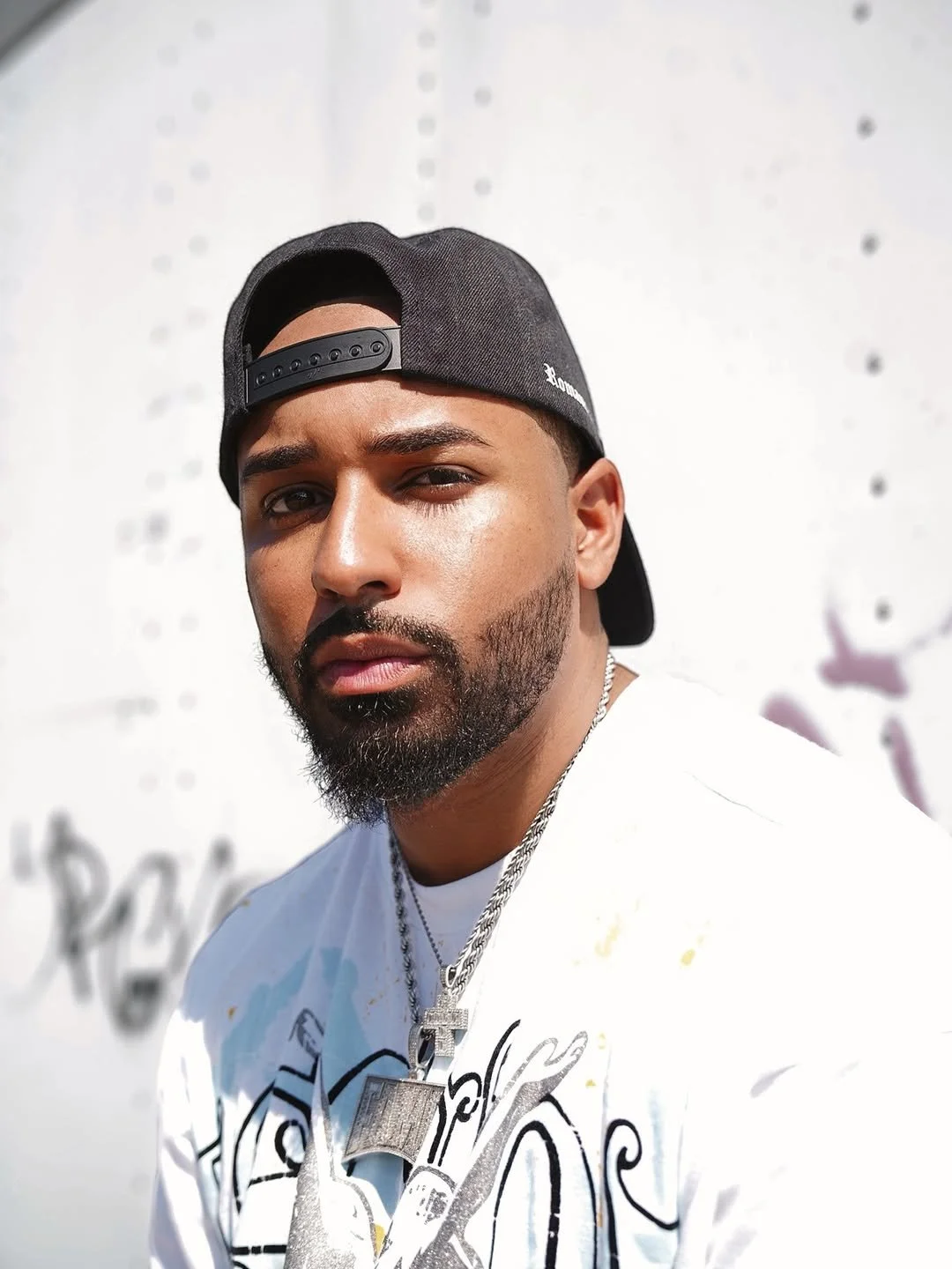 Fame Holiday with a beard and dark hair wearing a black cap backwards, a white graphic T-shirt, and silver chains, standing in front of a white wall with graffiti.