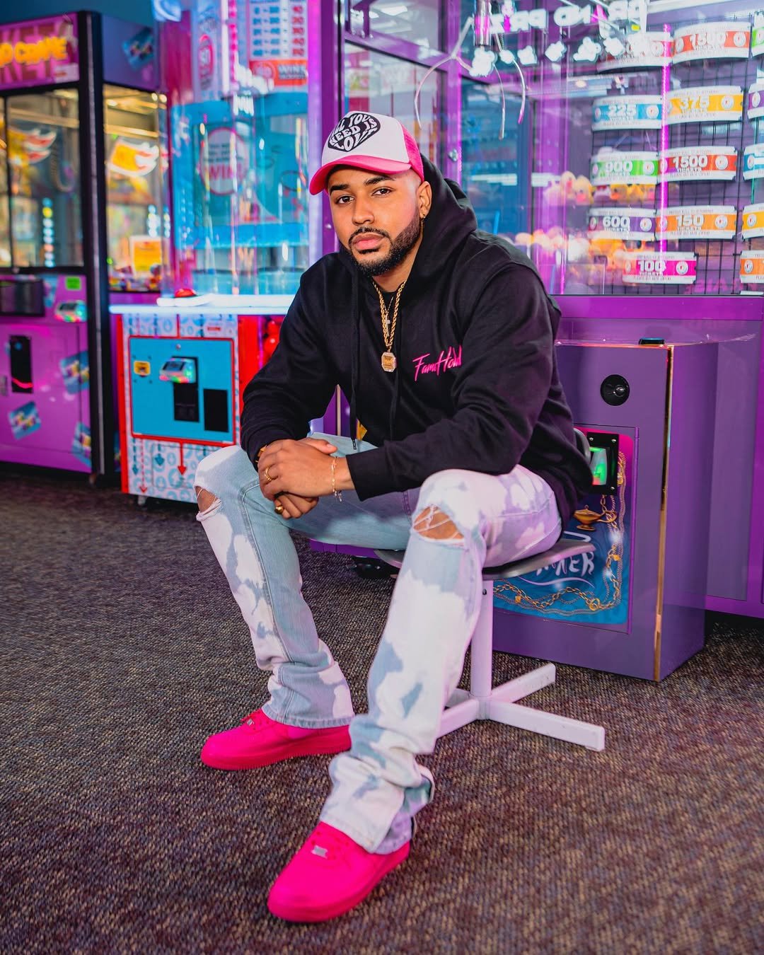 Fame Holiday sitting in arcade with purple neon lighting, wearing a black hoodie with pink lettering, white ripped jeans, pink shoes, and a pink and white cap. He has gold chains and bracelets.