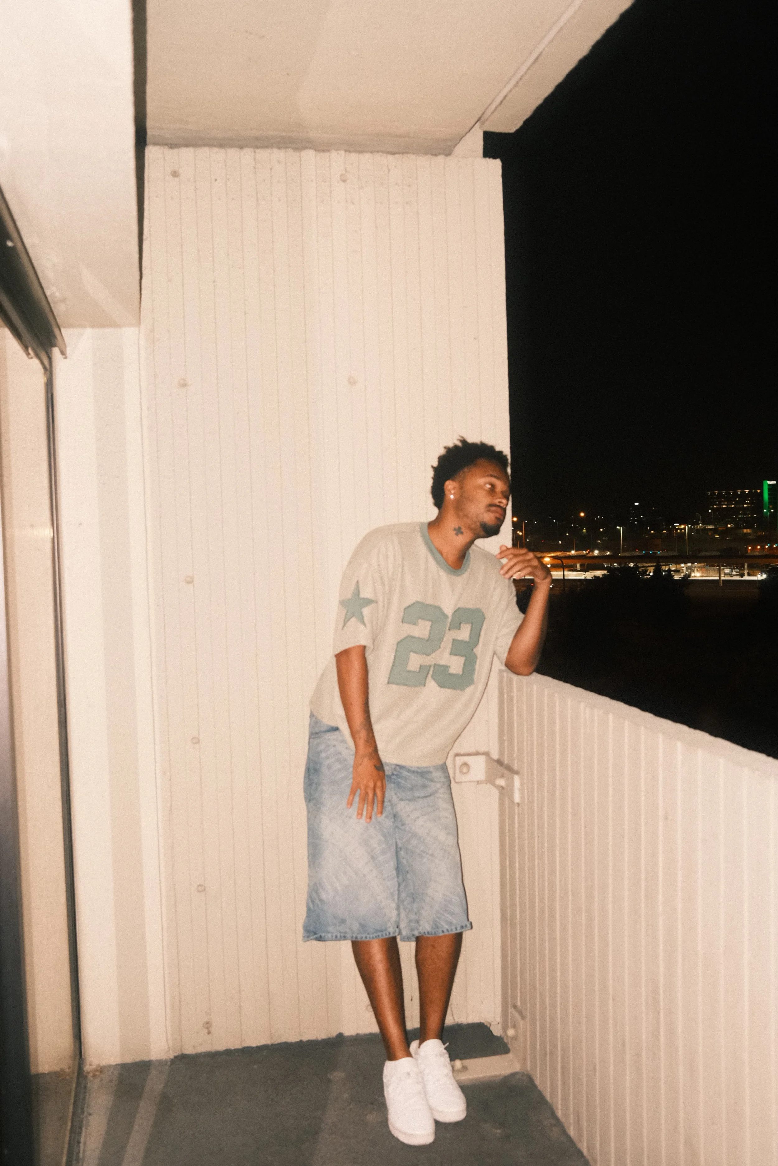 Jx.Zero on a balcony at night, wearing a gray sports jersey with the number 23, a denim skirt, and white sneakers, with a cityscape and dark sky in the background.