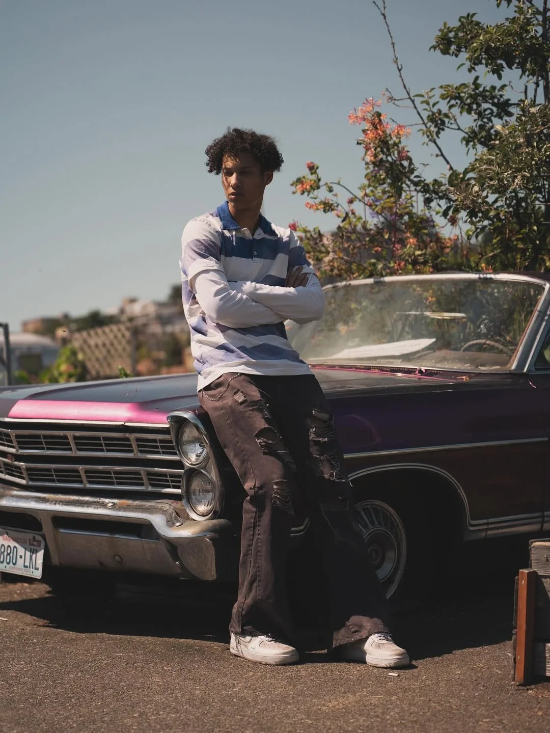 Jalen Jordan with curly hair wearing a striped long-sleeve shirt and ripped jeans leaning against a vintage black and pink car, with trees and flowers in the background on a sunny day. Still Can't Get Enough Single cover art.