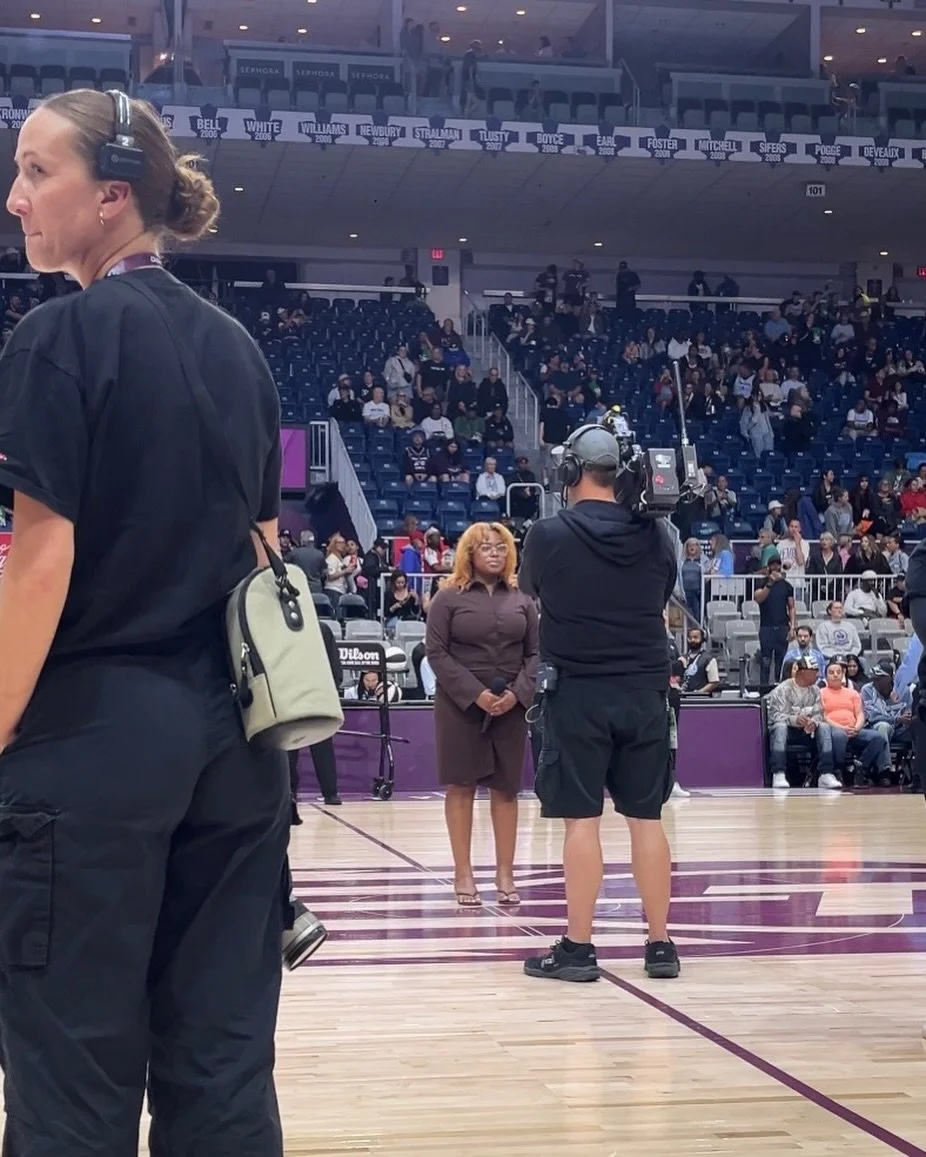  DĀHNI in a brown dress and glasses standing on a basketball court holding a microphone, being filmed by a cameraman. Several other people are seated or standing in the background with some wearing masks.