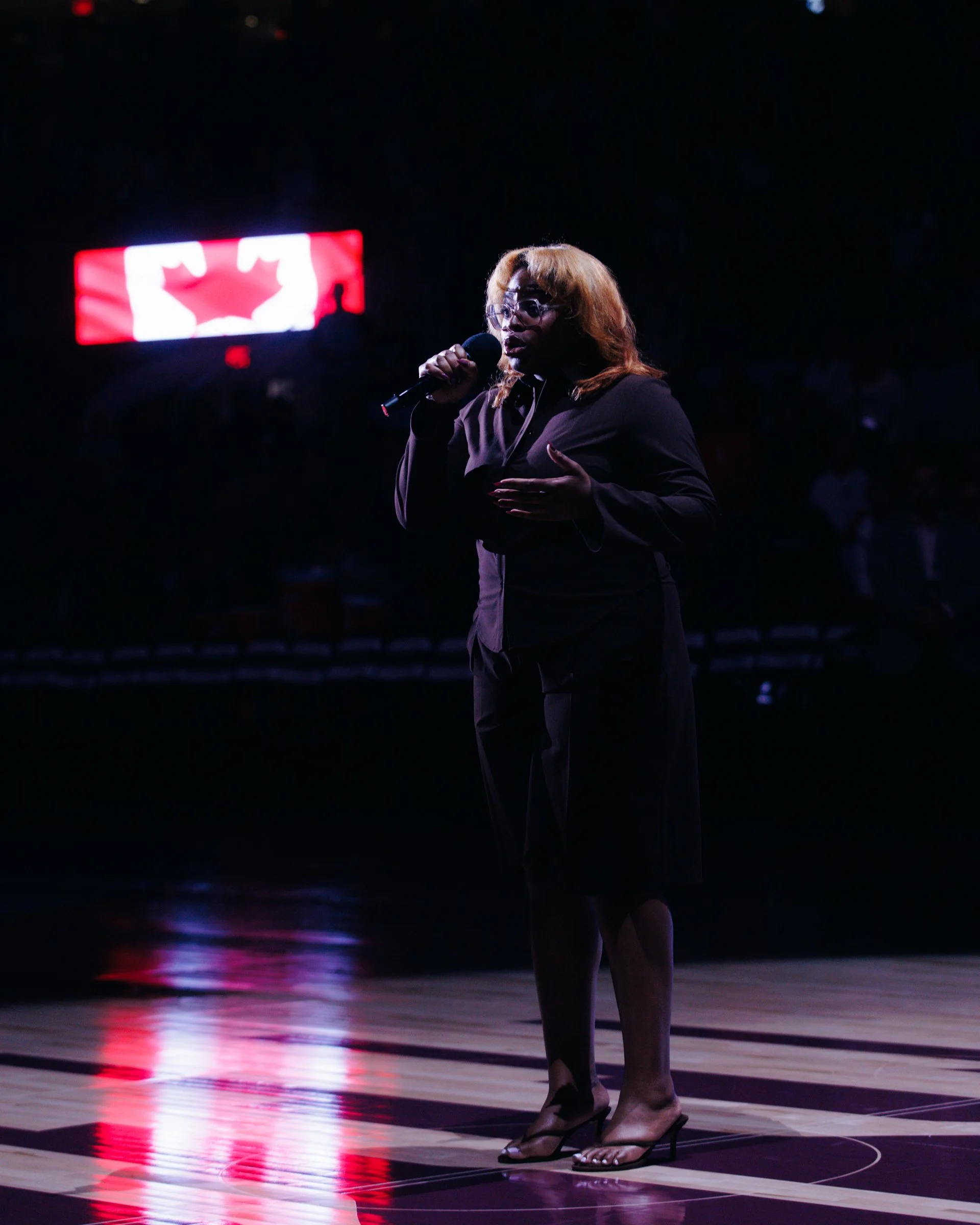  DĀHNI with long, blonde hair, wearing glasses, a dark blazer, and high heels, standing on a basketball court at night, holding a microphone, with stadium lights and a large digital screen displaying a red and white logo or image in the background.