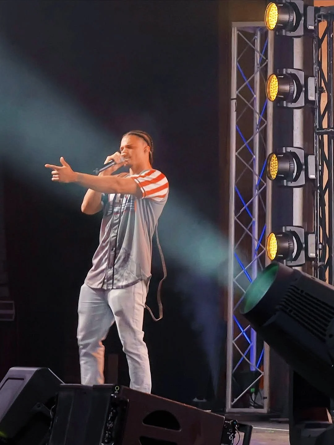 Deyton Jackson performing with braided hair holding a microphone and singing on stage, wearing a striped T-shirt and white pants, with stage lighting and equipment in the background.