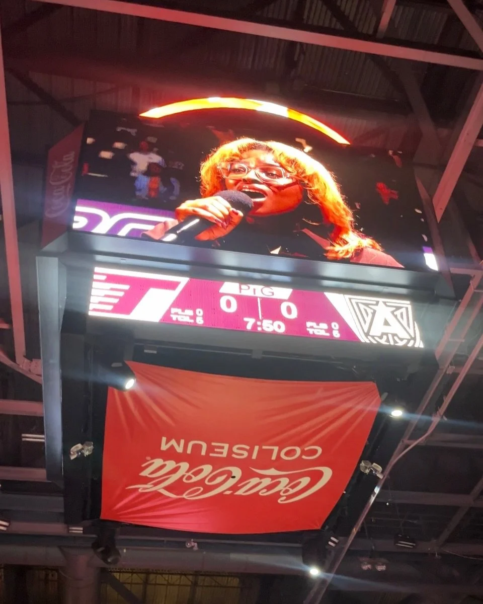  DĀHNI holding a microphone on a large sports scoreboard in a stadium, with Coca-Cola signage below.