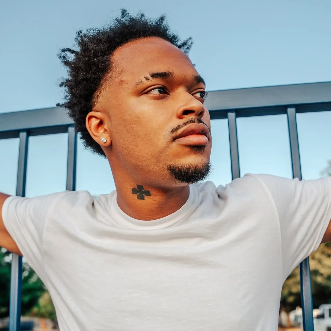 Jx.Zero with short, curly black hair and a small goatee, wearing a white t-shirt and a diamond earring, poses outdoors against a black metal fence during the daytime.