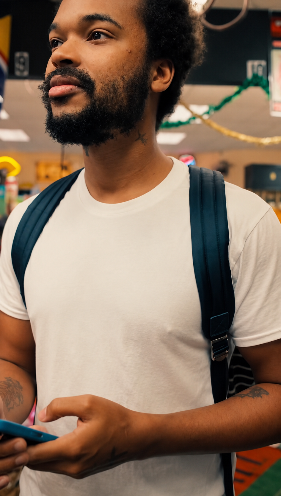 Jx.Zero with a beard and tattoos wearing a white t-shirt and a navy backpack in a store, holding a smartphone.