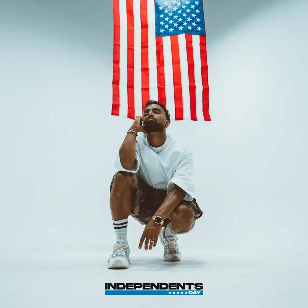 Futuristic in casual clothing squats in front of an American flag hanging on a plain white wall, with a logo at the bottom that reads 'Independents Day' and five blue stars.