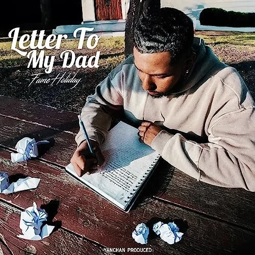 Fame Holiday writing a letter to his dad in a notebook, sitting at a wooden table outdoors with crumpled papers around him, on a sunny day.