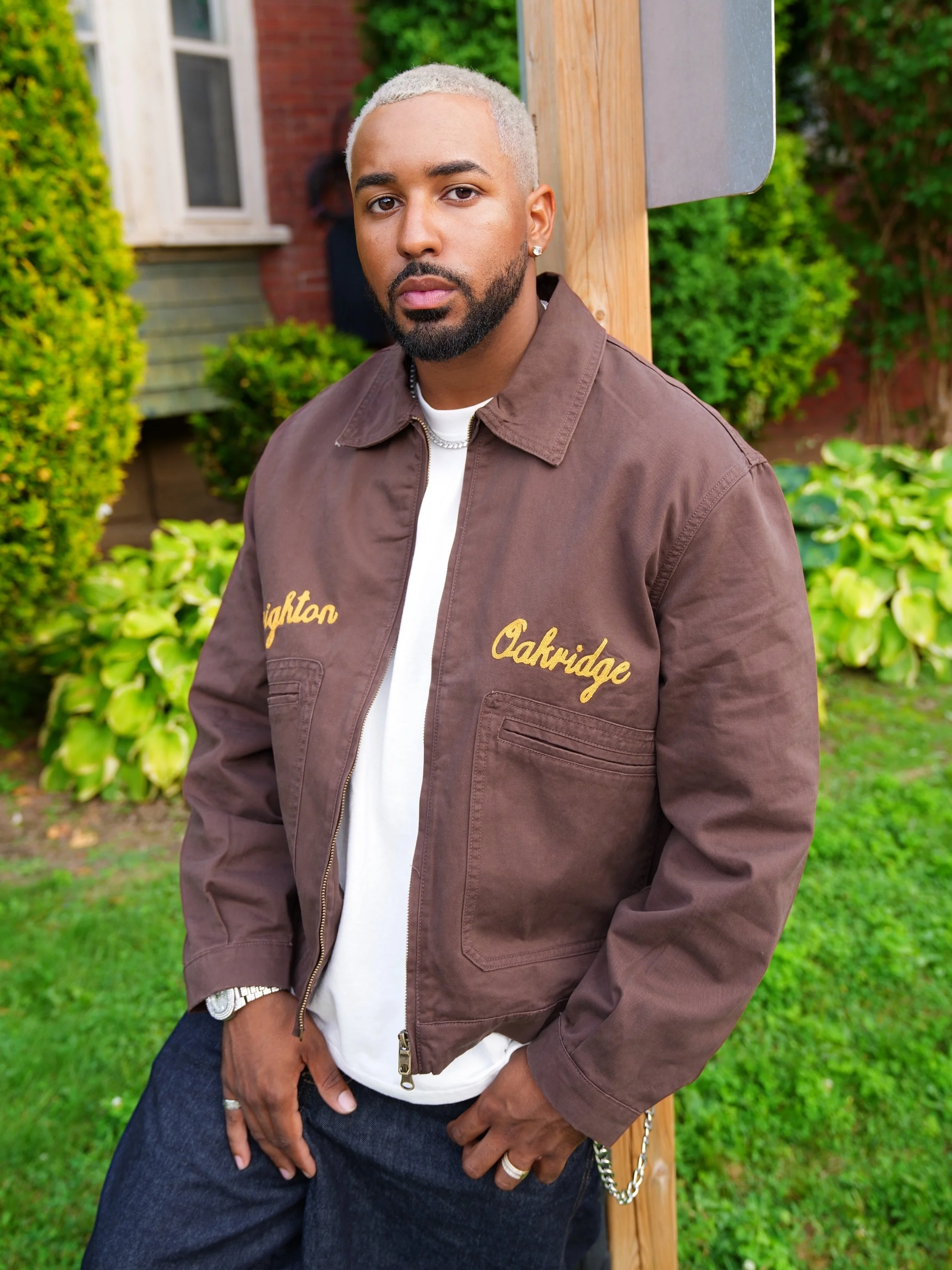 Fame Holiday with blonde hair, a beard, and earrings standing outdoors near a wooden post and greenery, wearing a brown jacket with gold embroidery and a white shirt underneath.