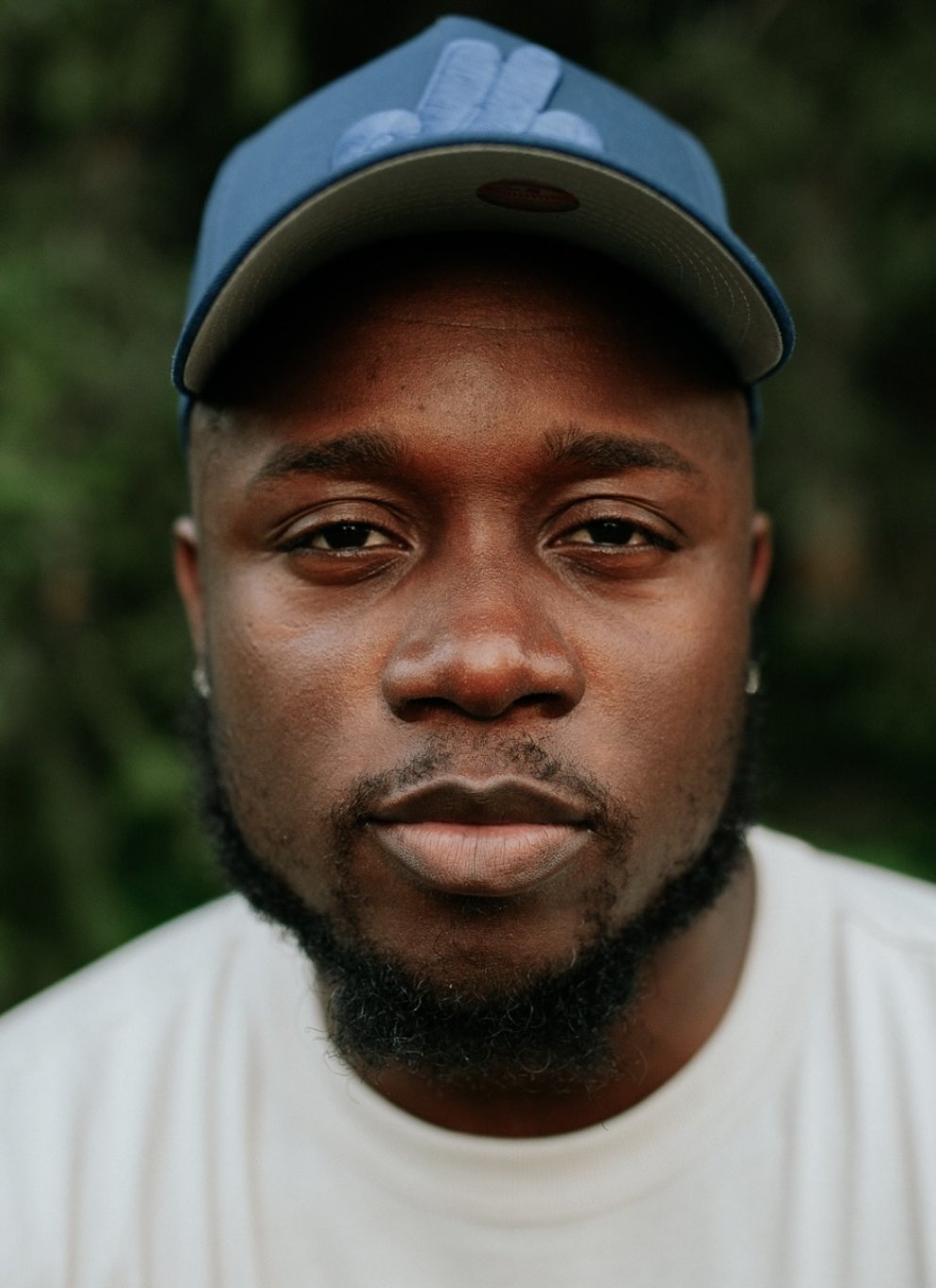 Close-up portrait of Kojo Dave wearing a blue cap and a white shirt, with a slight smile, dark skin, and a well-groomed beard, outdoors with blurred greenery in the background.