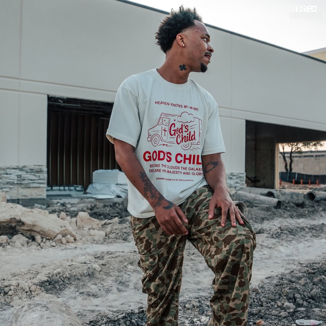Jx.Zero with tattoos, wearing a white graphic t-shirt and camo pants, stands outdoors on a construction site with dirt and rocks. Behind him is a modern building under construction.