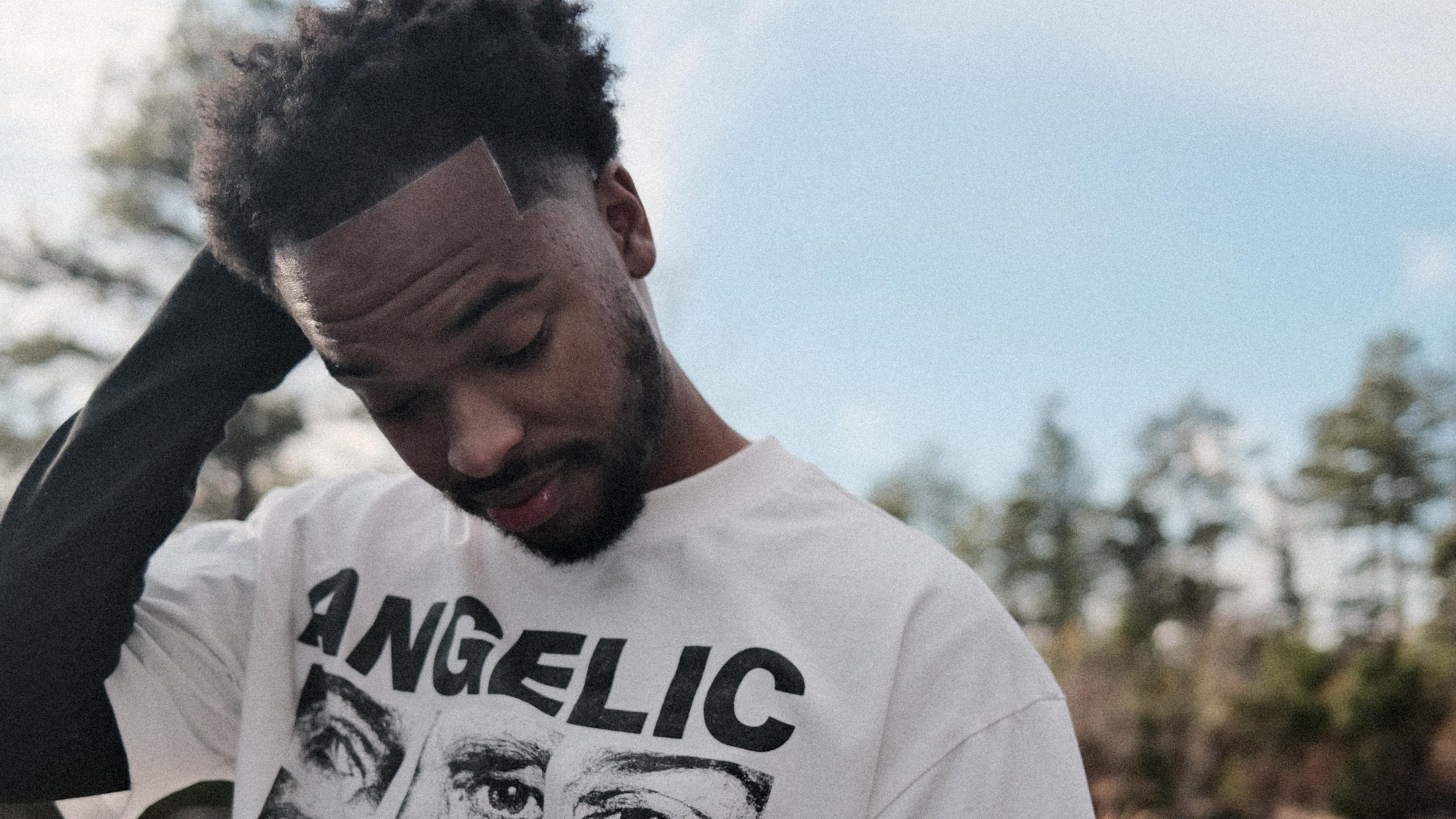 Jx.Zero with curly dark hair, a beard, and a bandage on his forehead, wearing a white T-shirt with a graphic print, is outdoors with trees and a cloudy sky in the background. He is touching the back of his head and looking down.
