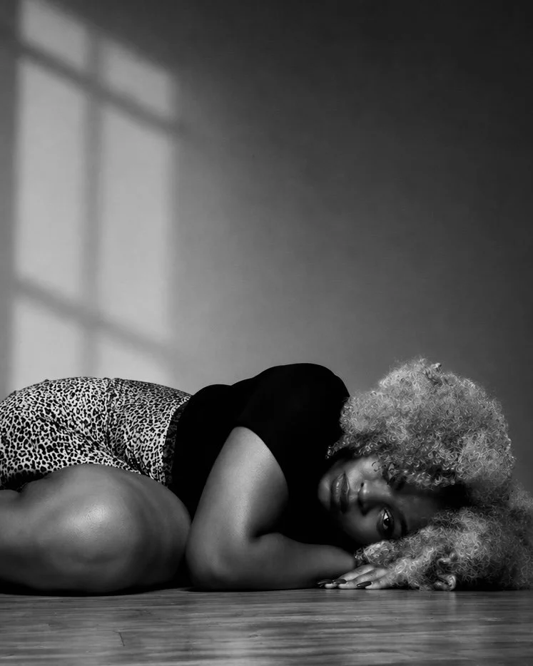 DĀHNI with curly blonde hair lying on the wooden floor, resting her head on her arm and gazing at the camera, with a shadow of a window cast on the wall behind her.