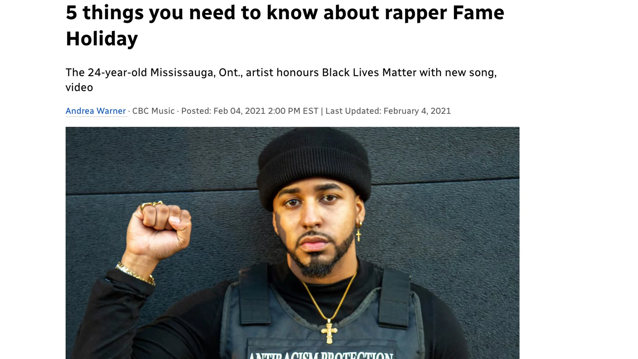 Fame Holiday with a serious expression, wearing a black beanie, gold jewelry, and a black shirt with a bulletproof vest, raising his right fist in front of a dark textured wall.