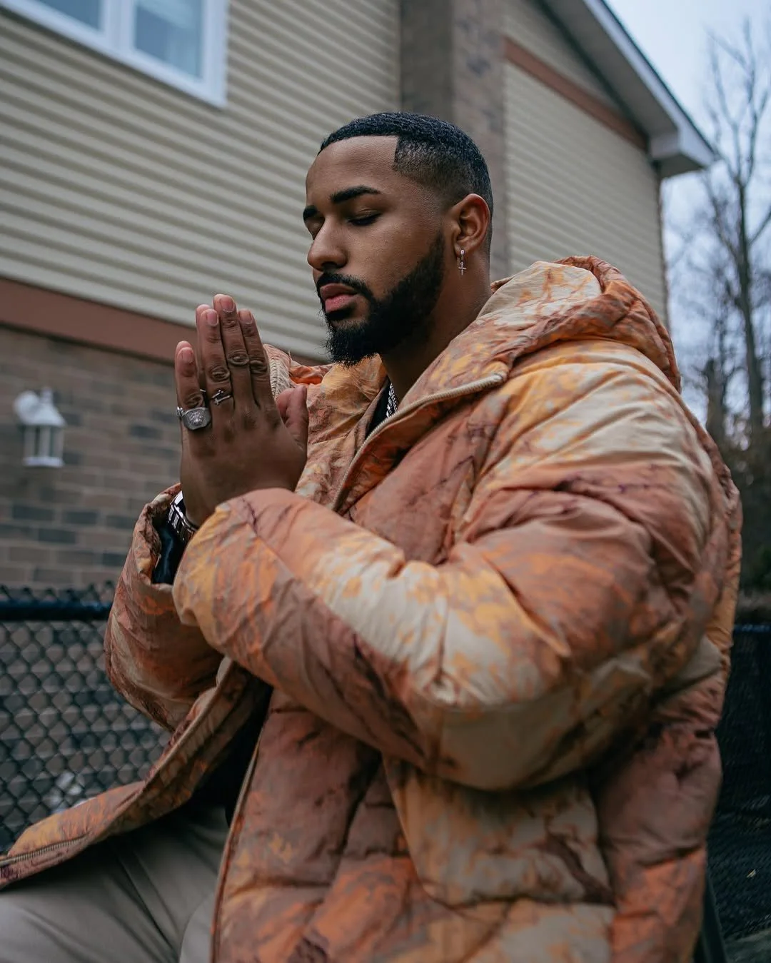 Fame Holiday with a beard, earrings, and rings, wearing an orange patterned jacket, sits outdoors with his eyes closed and hands pressed together in prayer or meditation.