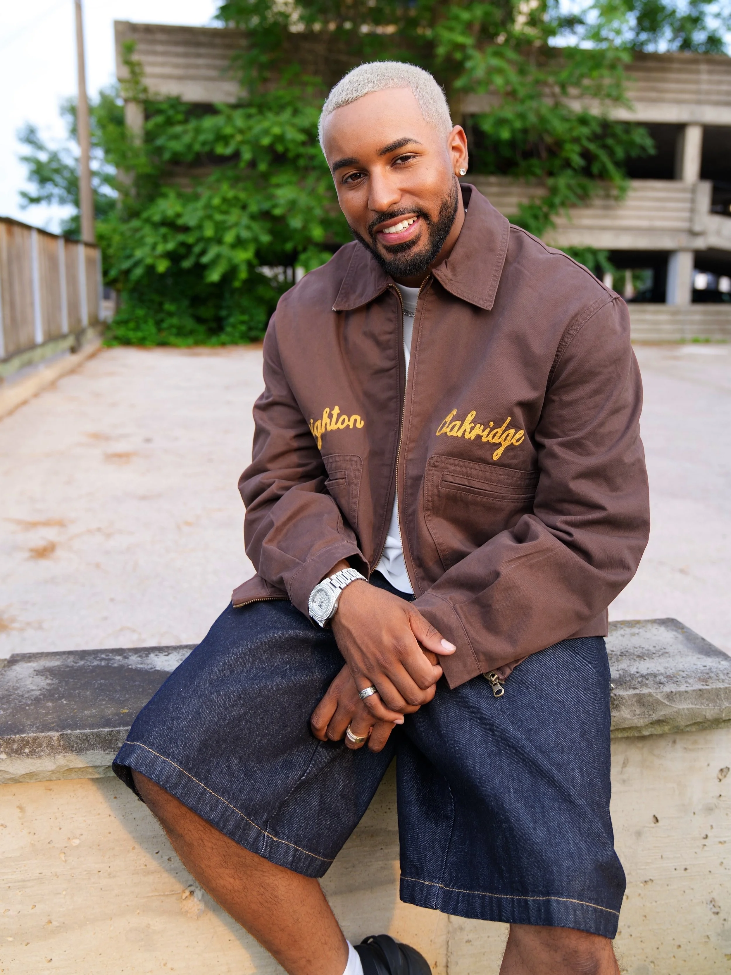 Fame Holiday with blonde hair, a beard, wearing a brown jacket, dark blue shorts, and black shoes, sitting outdoors on a low concrete wall with greenery and a wooden structure in the background.
