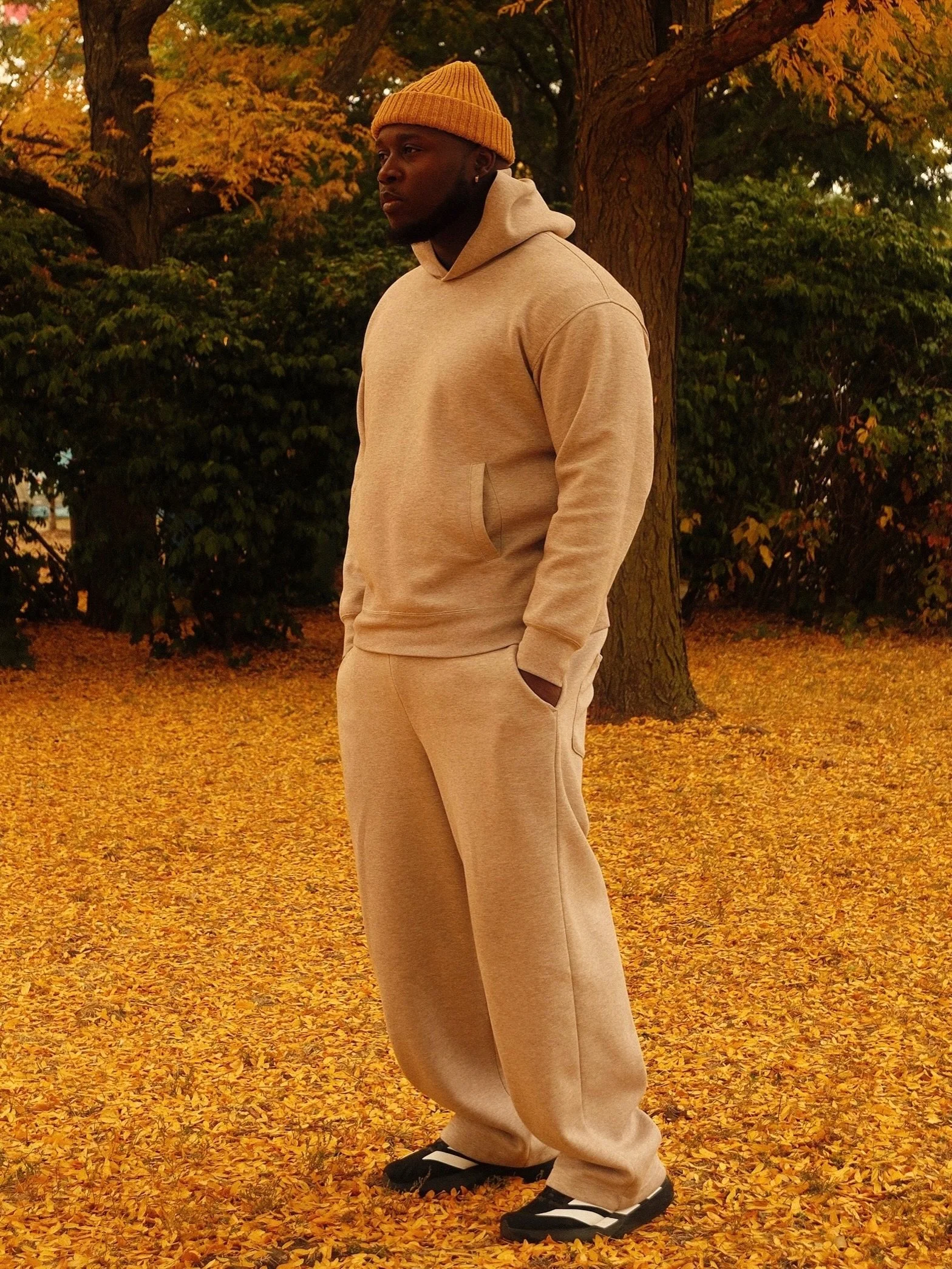 Kojo Dave in a beige hoodie and sweatpants standing outdoors on a bed of fallen yellow leaves during fall, with trees in the background.