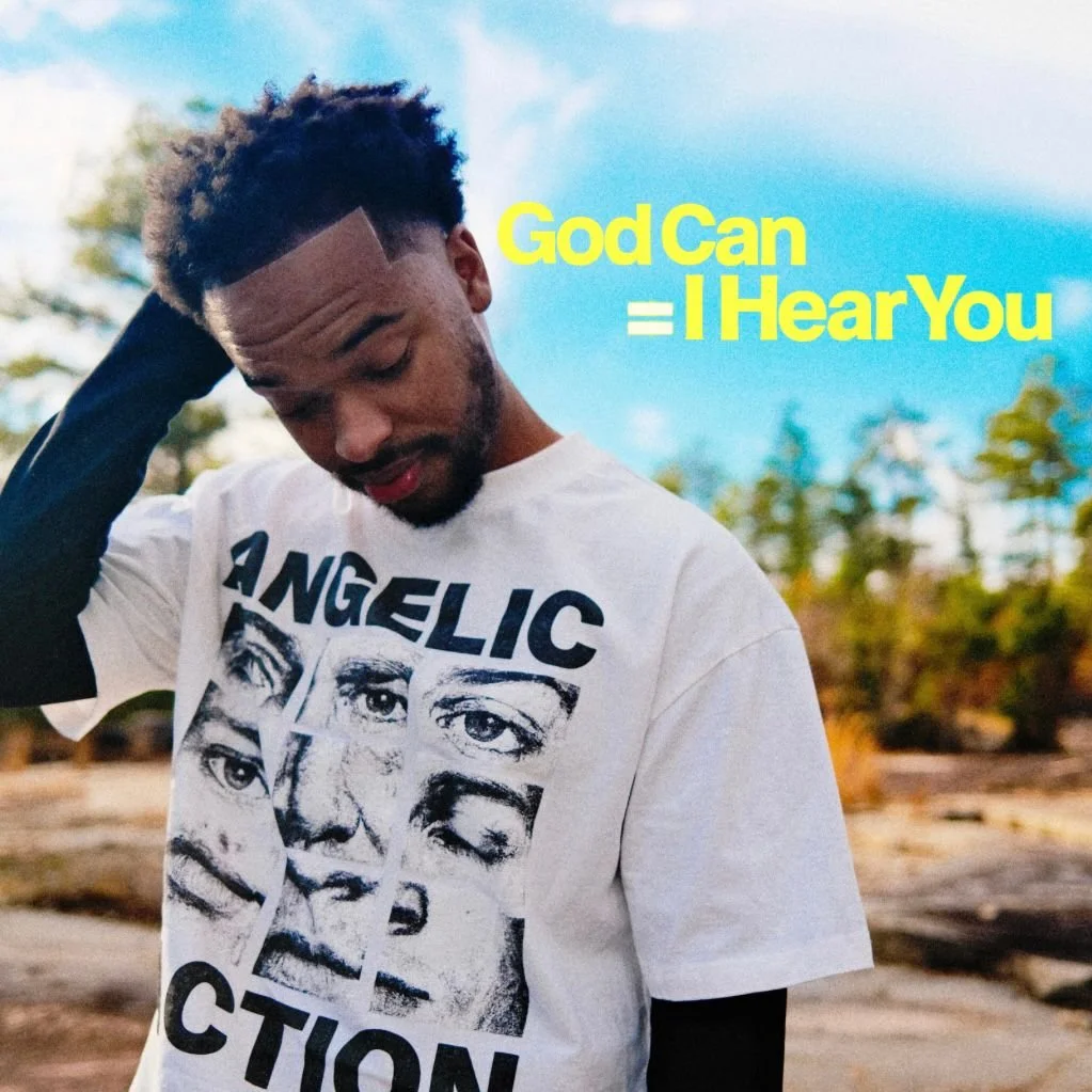 Jx.Zero with short curly hair and a beard, standing outdoors with trees and a blue sky in the background, wearing a white T-shirt with black and white printed faces and the word 'ANGELIC' on it. There is yellow text that reads 'God Can = I Hear You' 